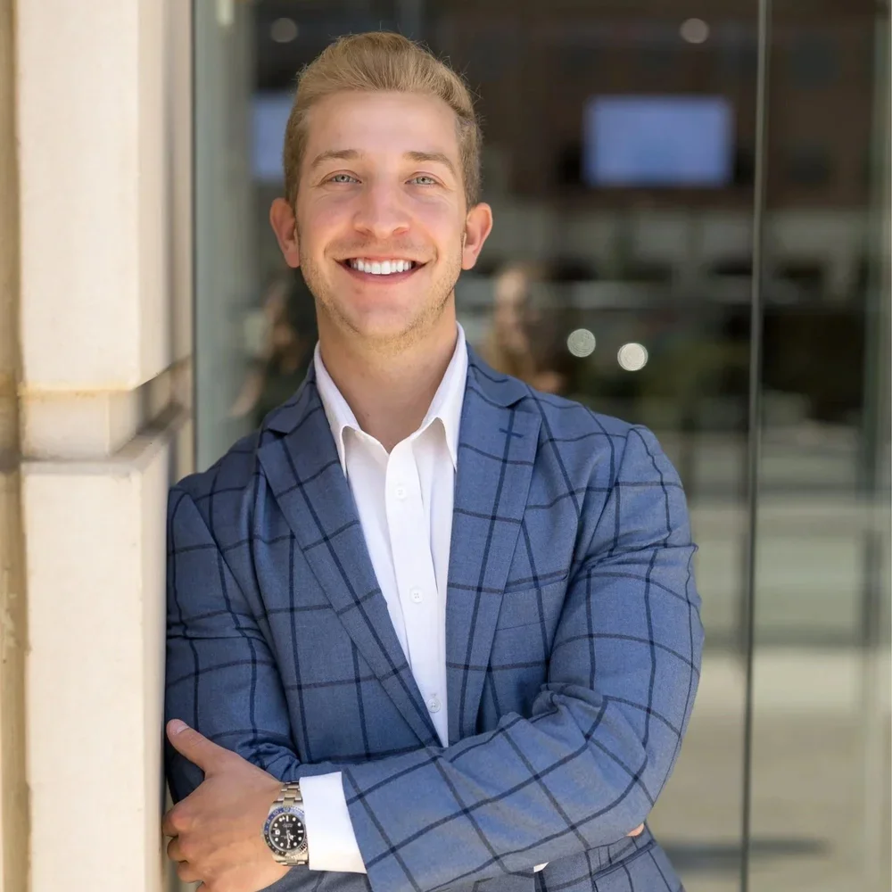 Young man in a blue checkered suit smiling and leaning against a wall near a glass window.