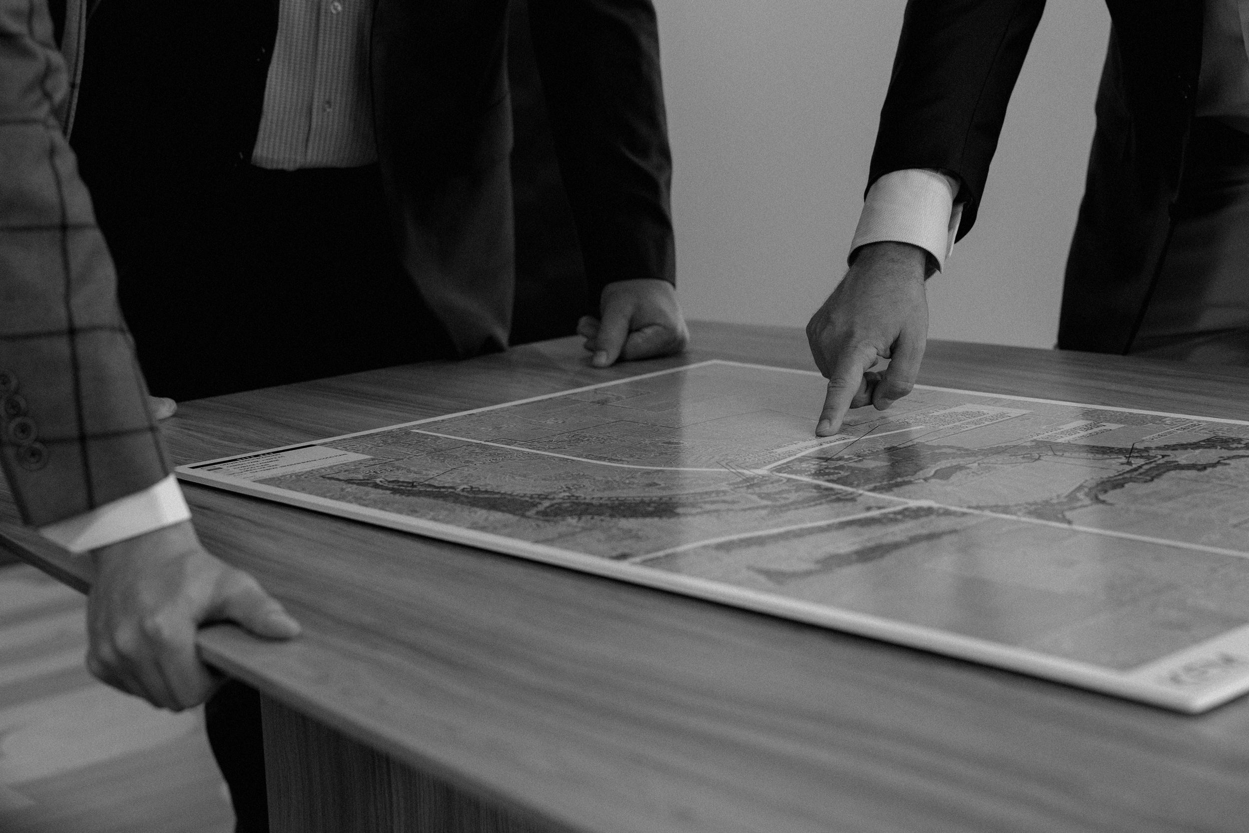 Several people in business attire are gathered around a wooden table, examining and discussing documents or maps laid out on the surface.