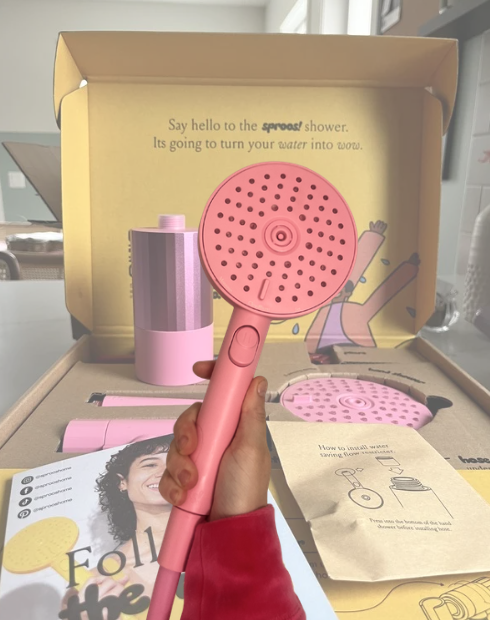 A pink showerhead held by a person's hand in front of a yellow box containing the same showerhead, instructional materials, and a small pink bottle.