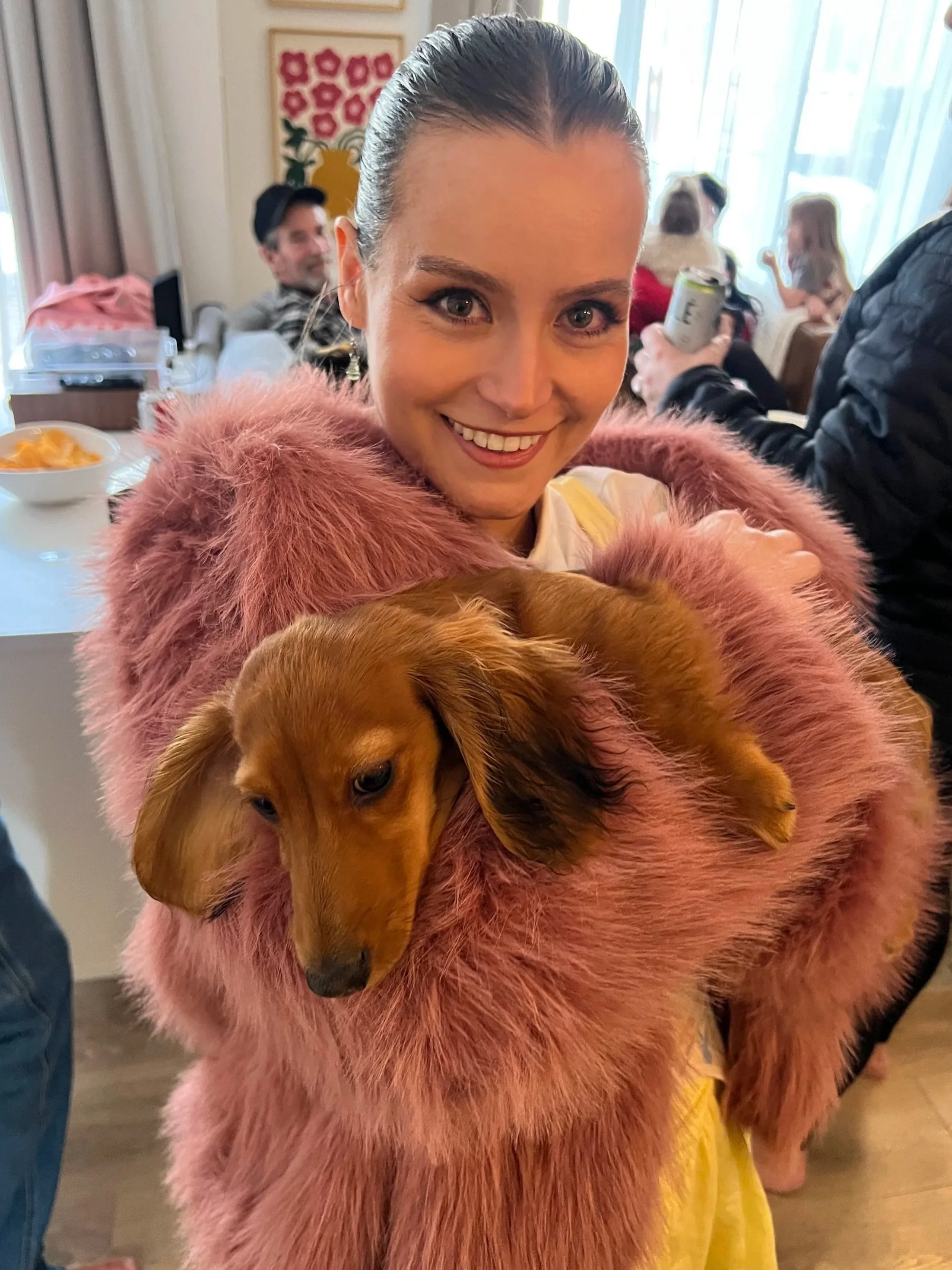 A woman with brown hair and a pink fur coat is holding a small, long-haired dachshund puppy. The woman is smiling and standing indoors with a group of people in the background.