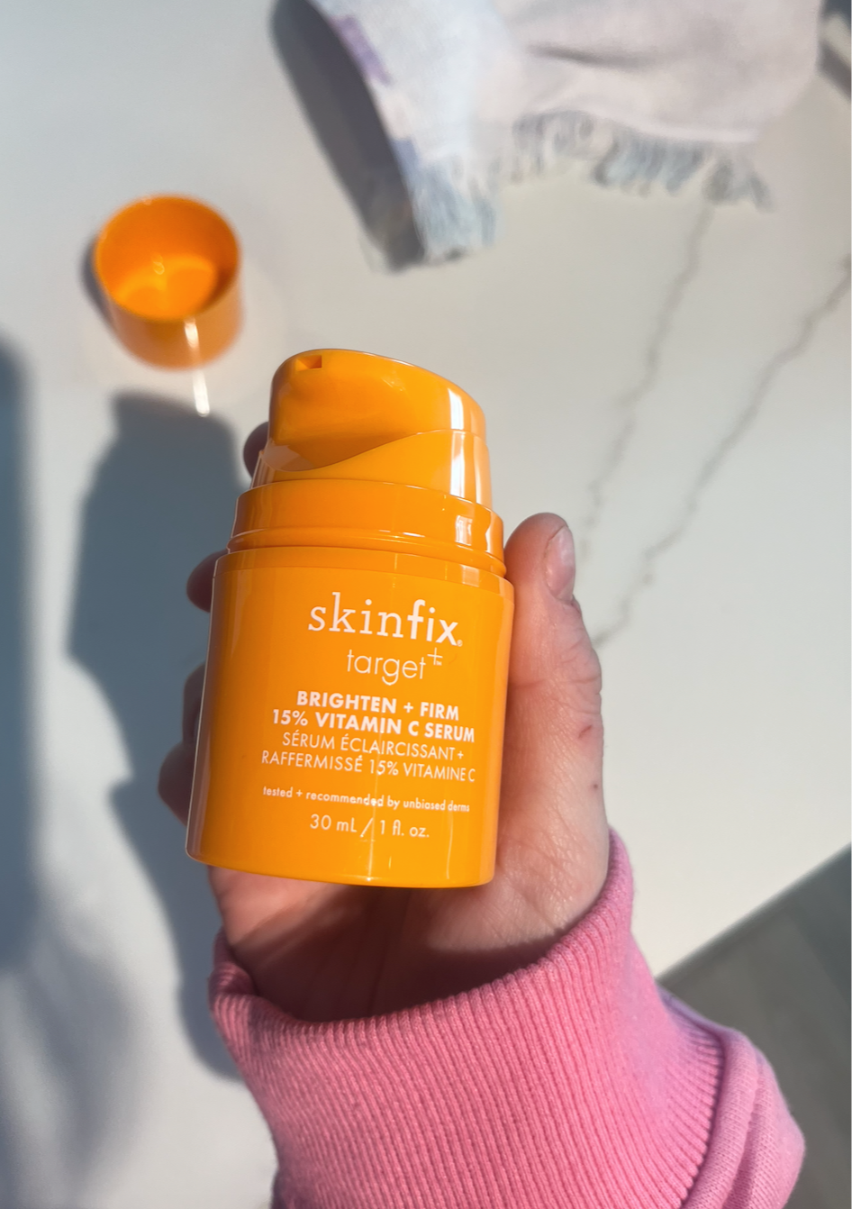A hand holding an orange bottle of SkinFix target brightening and firm 15% Vitamin C serum on a white surface, with an open orange cap and an orange container in the background.
