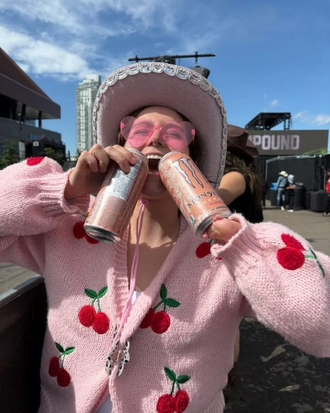 Woman wearing a pink cherry-patterned sweater, pink sunglasses, and a large white brimmed hat, smiling and holding two cans of Monster energy drinks near her open mouth outdoors on a sunny day.