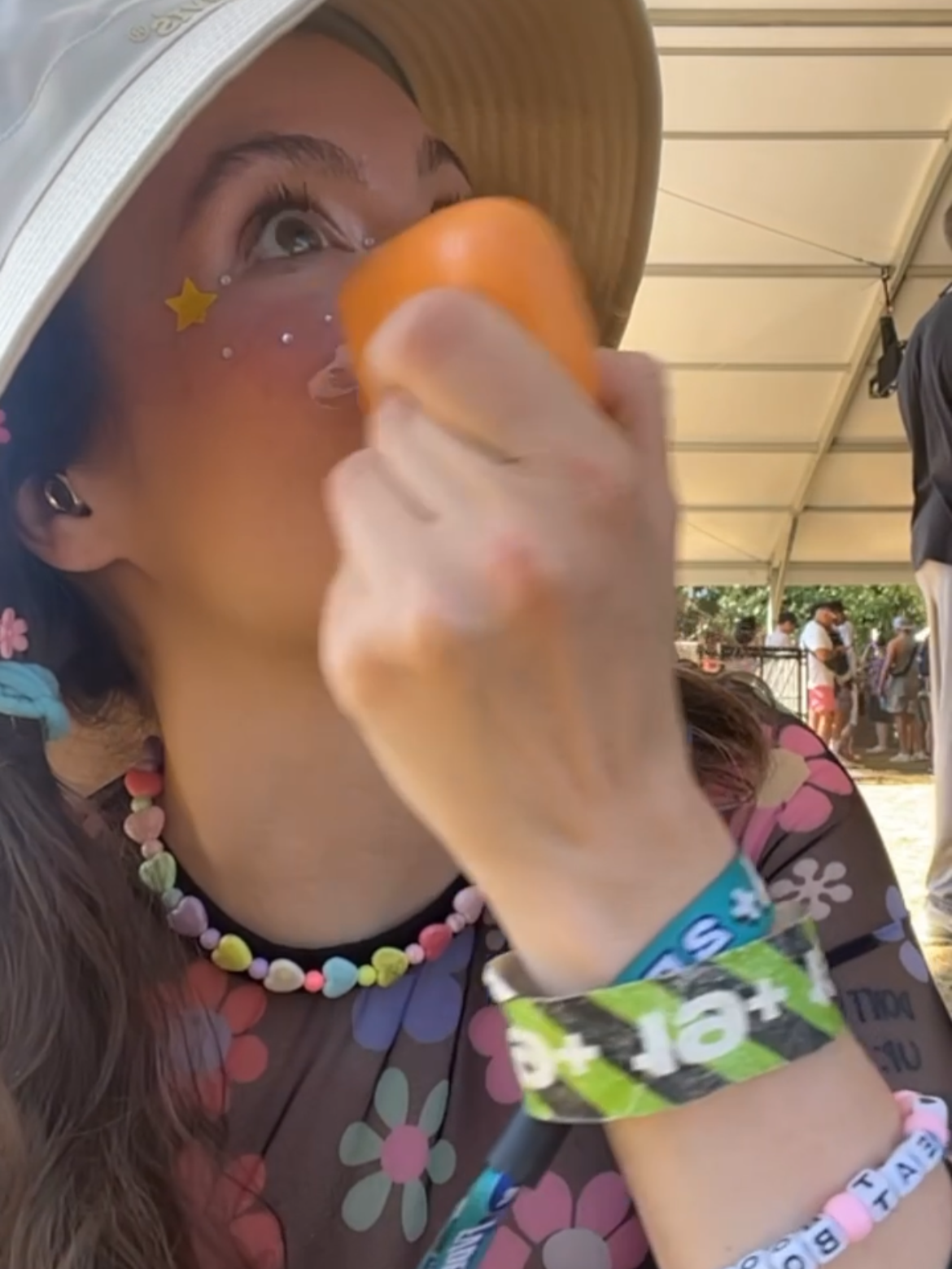 A person wearing a straw hat, colorful beads necklace, and Festival wristbands is drinking from an orange cup at an outdoor event under a tent.