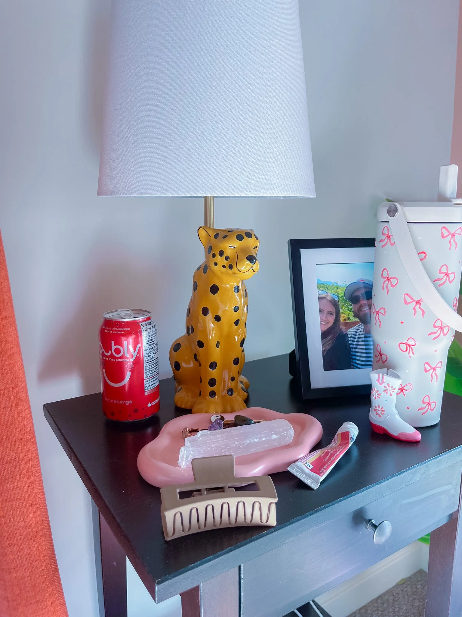 A black side table with a yellow leopard-shaped lamp, a framed photo of a smiling couple, a can of a beverage, a pink tray with a selenite crystal, some small stones, and a nail clipper, and a white bag with pink bows and a small white boot figurine with pink accents.