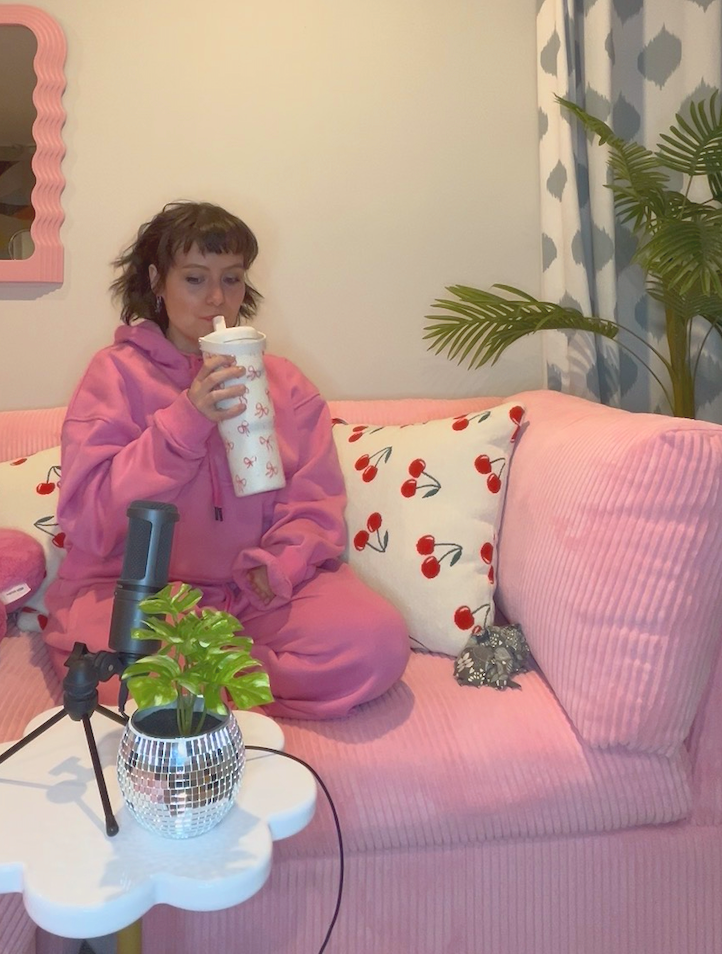 A woman in a pink hoodie and matching pants sitting on a pink couch, drinking from a white cup with a logo. There are cherry-printed pillows on the couch, a small white coffee table with a green plant in a decorative pot, a microphone on a stand positioned near the table, and large green plants in the background. Curtains with a diamond pattern are also visible.