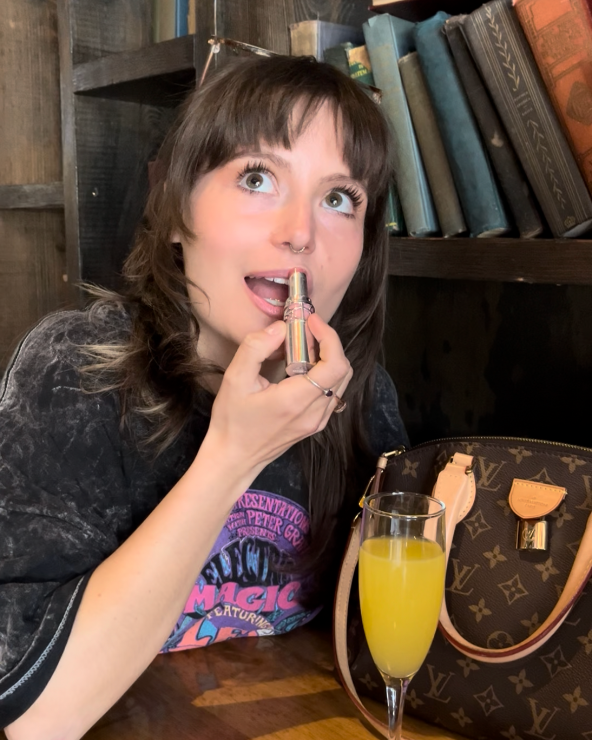 A woman with brown hair and a septum piercing applying lipstick at a table, with a yellow drink and a Louis Vuitton bag nearby, in a setting with bookshelves in the background.