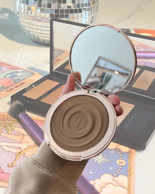 Hand holding open compact mirror with pressed powder inside, surrounded by makeup items such as eyeshadow palettes and brushes, with decorative items and colorful illustrations in the background.