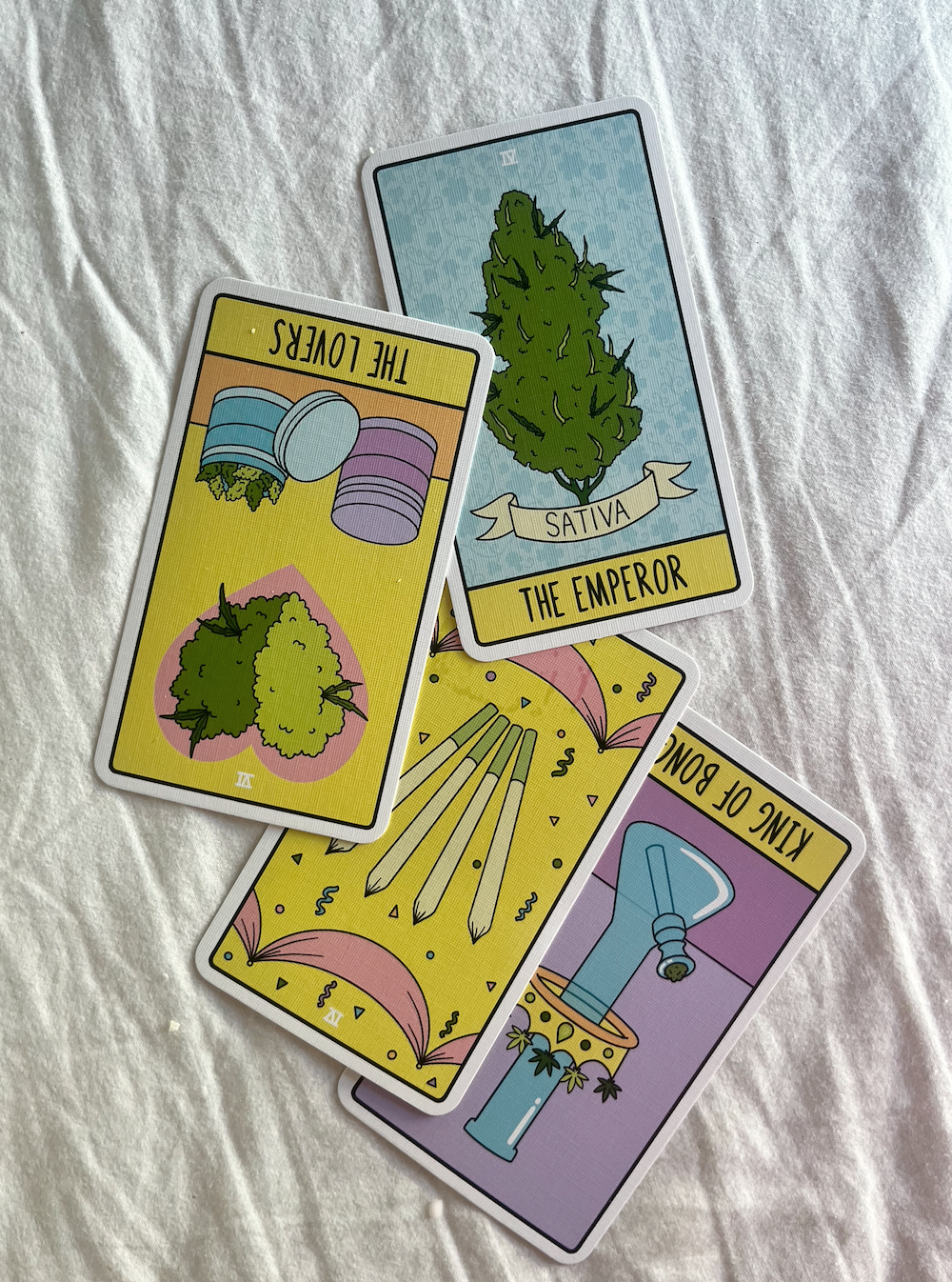 A set of four tarot cards laid out on a white fabric surface. The cards include The Emperor depicted as a large cannabis plant, King of Bongs showing a bong with plants, Three of Wands featuring three wands, and The Lovers showing two containers with plants.
