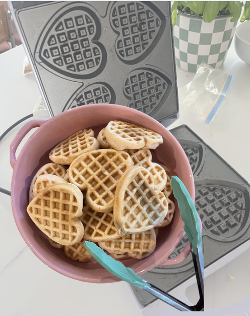 A pink bowl filled with waffle cookies, some heart-shaped, sits on a countertop with a waffle iron in the background and a potted plant nearby.
