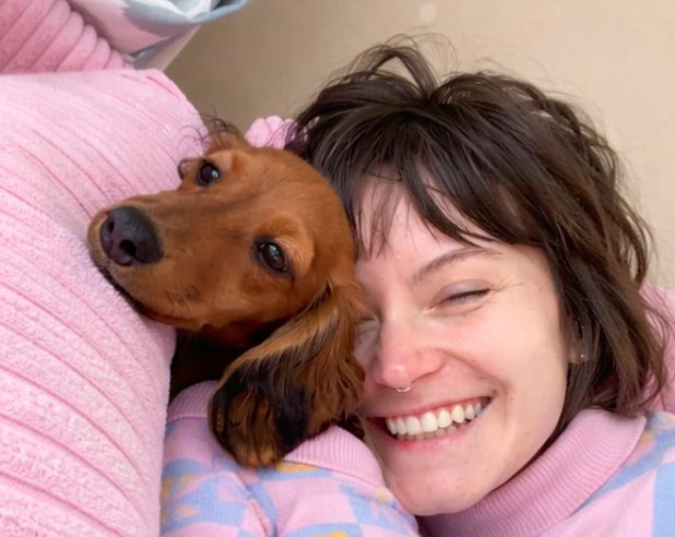 A woman with short, tousled brown hair and a septum piercing smiling with her eyes closed, lying closely with a brown dog with long ears resting its head on her shoulder.