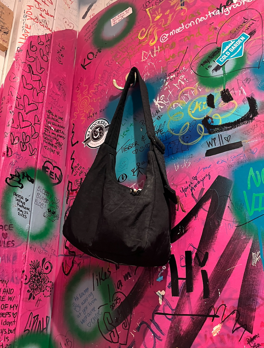 Black shoulder bag hanging on a colorful graffiti wall covered with handwritten messages and stickers.