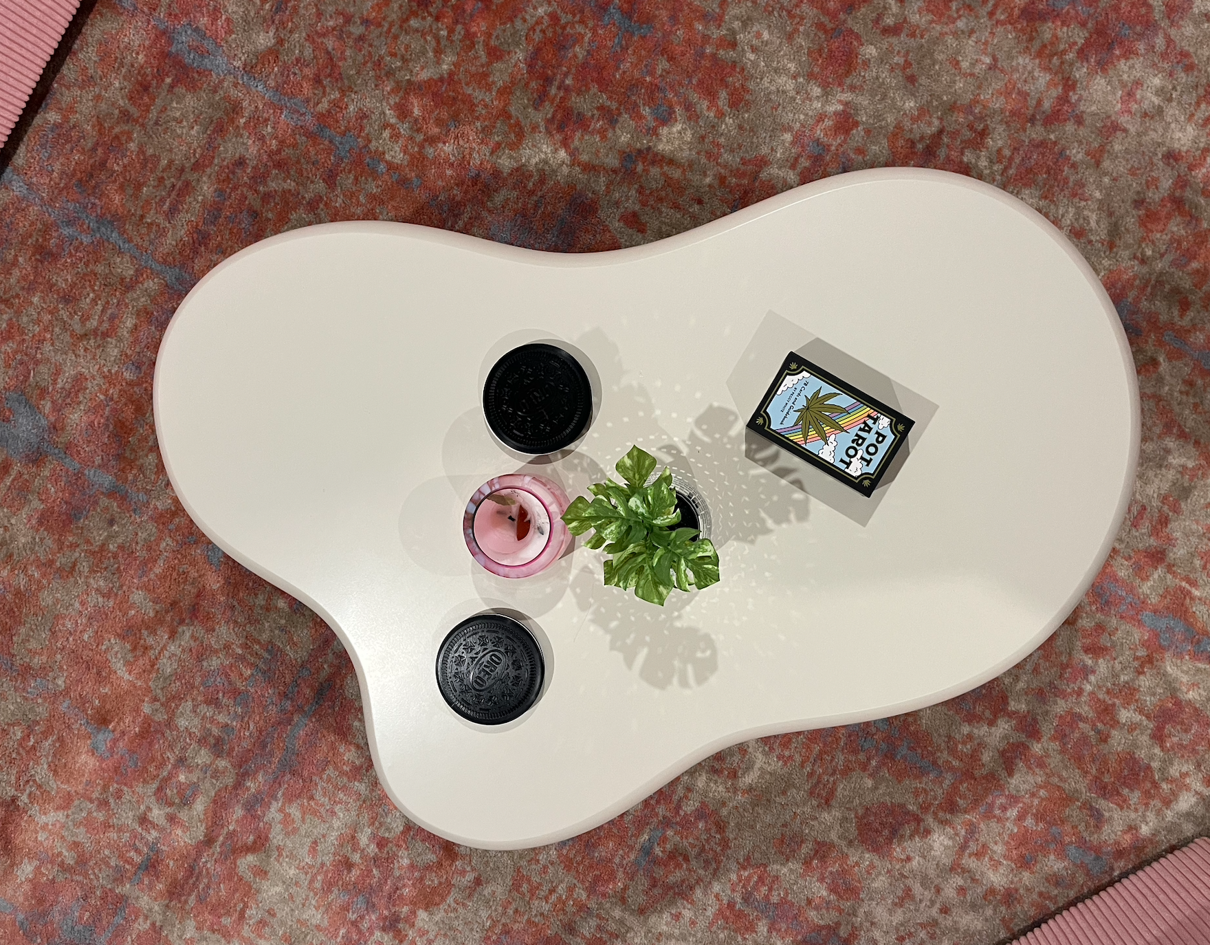 A white coffee table with irregular shape on a patterned rug, holding two black jars, a pink candle, a green potted plant, and a box with a colorful label depicting a marijuana leaf.