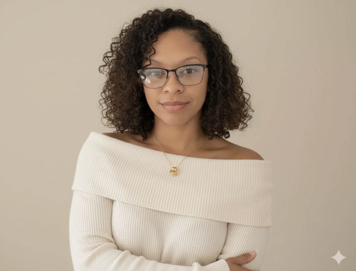 Young woman with curly hair and glasses wearing a cream off-shoulder sweater and a gold necklace, standing against a plain beige background.