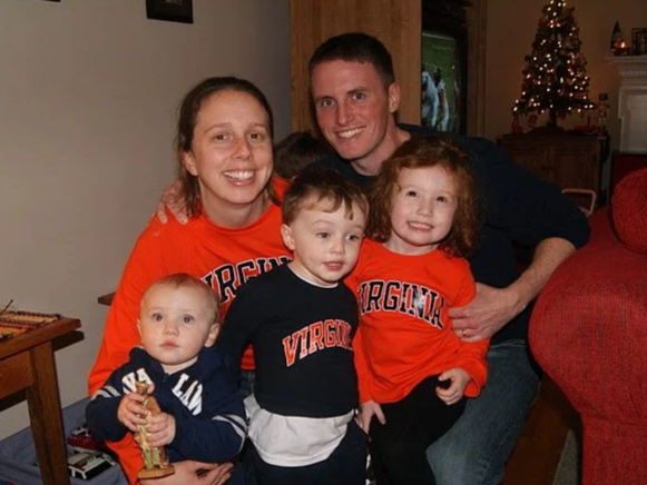 J.P., his wife Maureen, and their three young kids celebrating Christmas indoors, with a decorated tree in the background. The adults and children are smiling and wearing University of Virginia clothing.