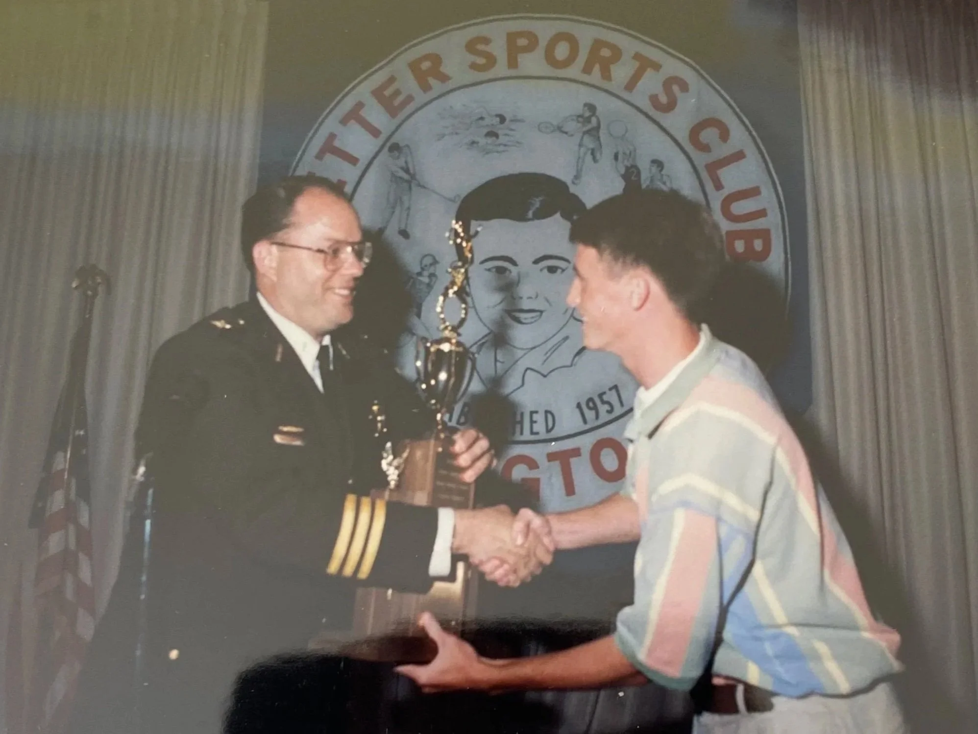 A man in a military uniform is holding a trophy and shaking hands with a young J.P. in a striped shirt during an award ceremony at Wetter Sports Club, with a large children’s illustration and the club's banner in the background.