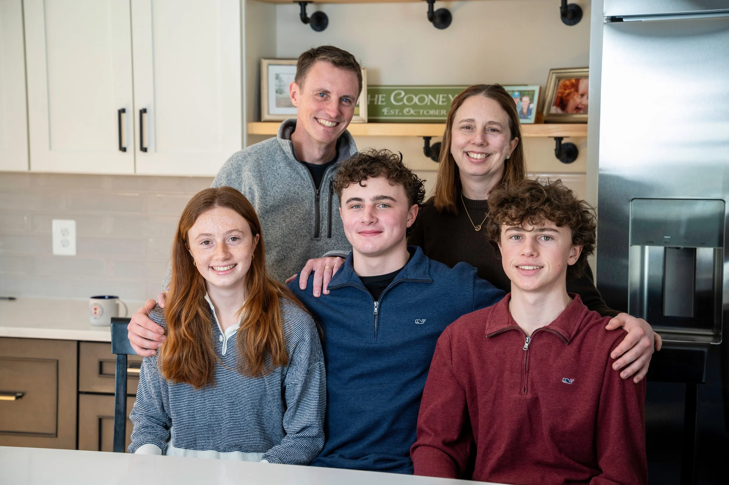 J.P.'s family smiling and posing for a photo in a kitchen. Two adults stand in the back, two teenagers sit in front, and one teenager is in the middle. The kitchen has light-colored cabinets, framed photographs, and a sign that reads 'The Cooneys'.