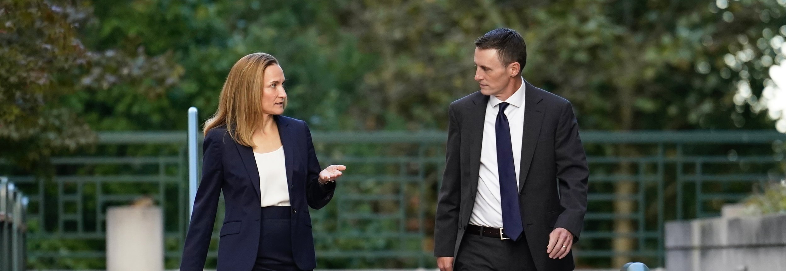 A man and woman in business attire are walking and talking outdoors, with trees and a fence in the background.
