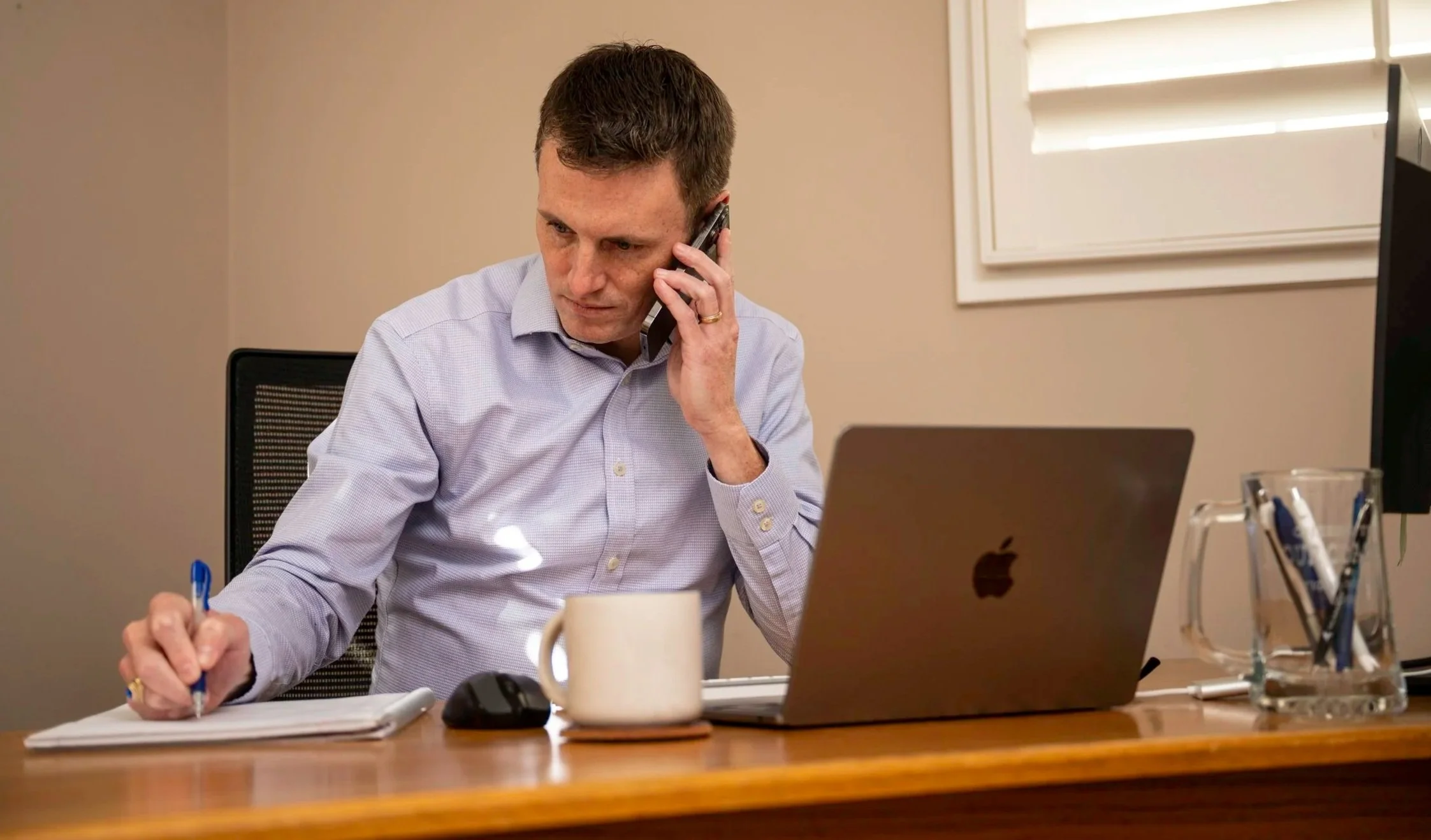 J.P. sitting at a desk, talking on the phone, writing on a notepad, with a laptop, coffee mug, and office supplies on the desk.