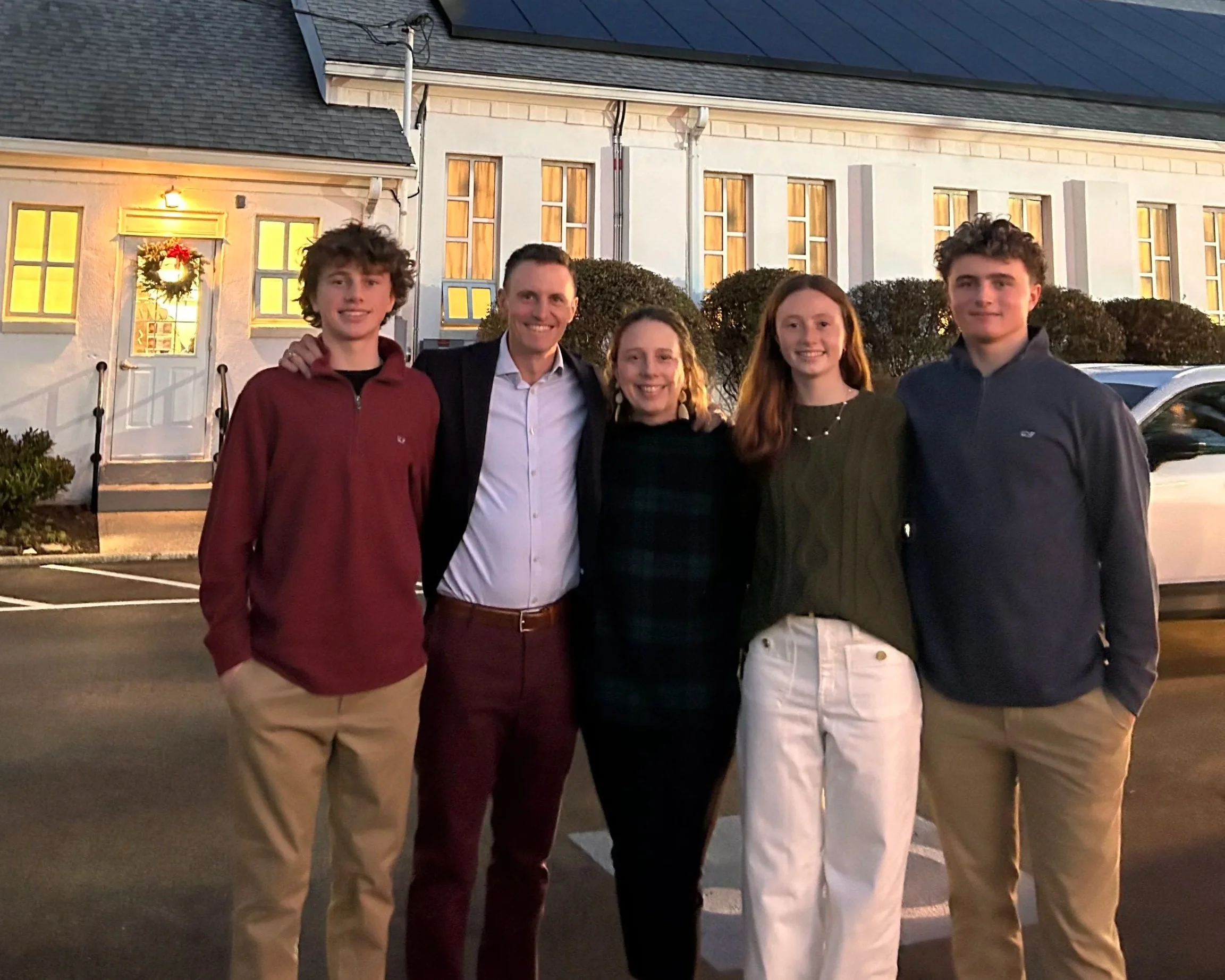 Group of five people standing outside in front of a building decorated for Christmas, smiling at the camera during sunset.