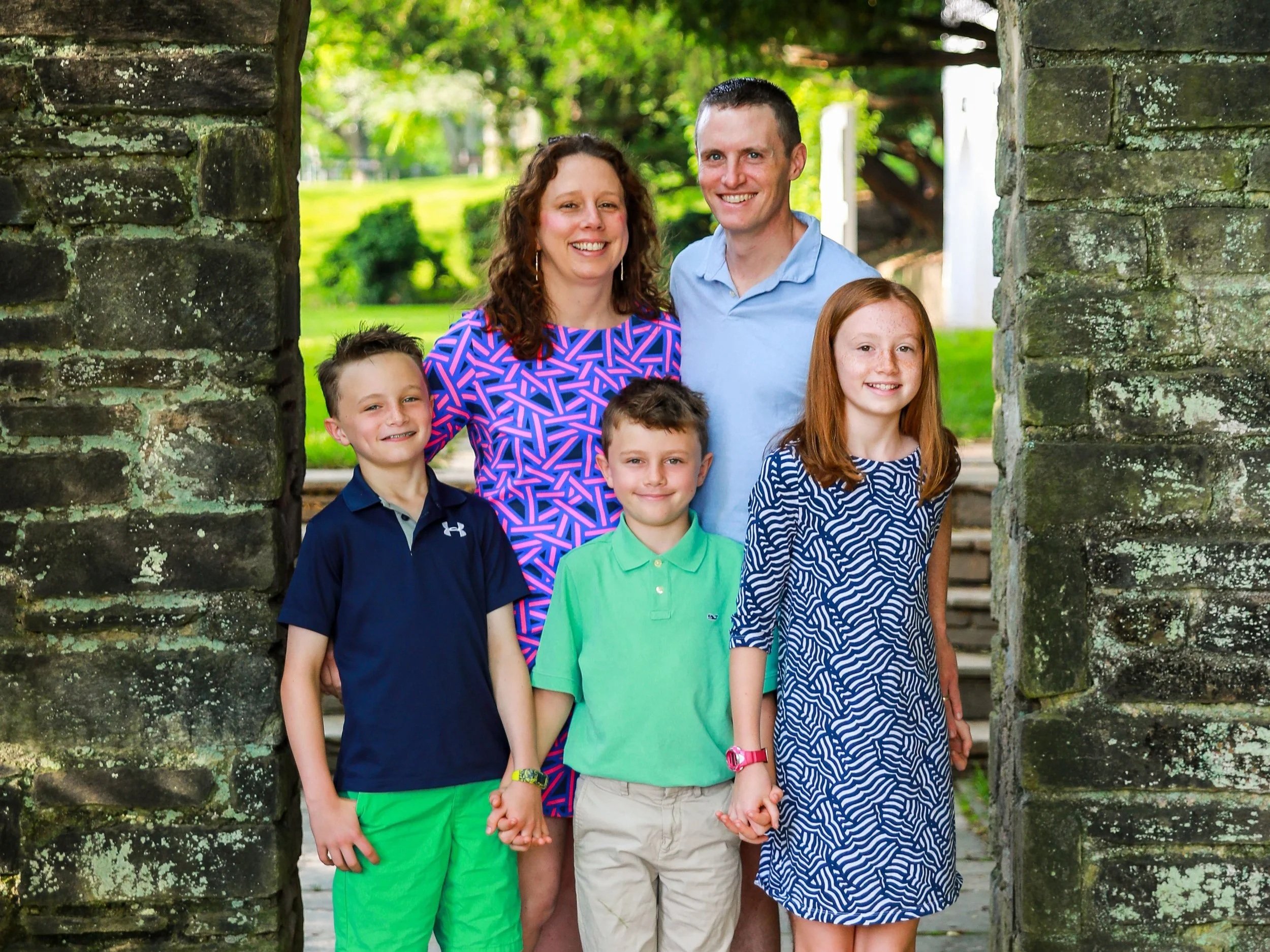 A family of six standing outdoors between stone pillars. The family includes two adults and four children. They are smiling and holding hands in a park with green trees in the background.