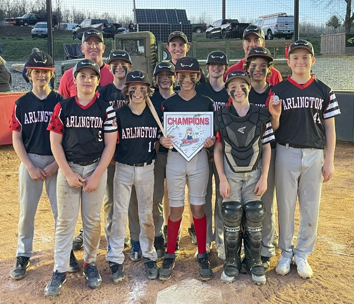 Little league baseball team in black and red uniforms with 'Arlington' on the shirts, posing on the field with a framed championship sign, some players wearing catcher's gear, and smiling coaches behind them.