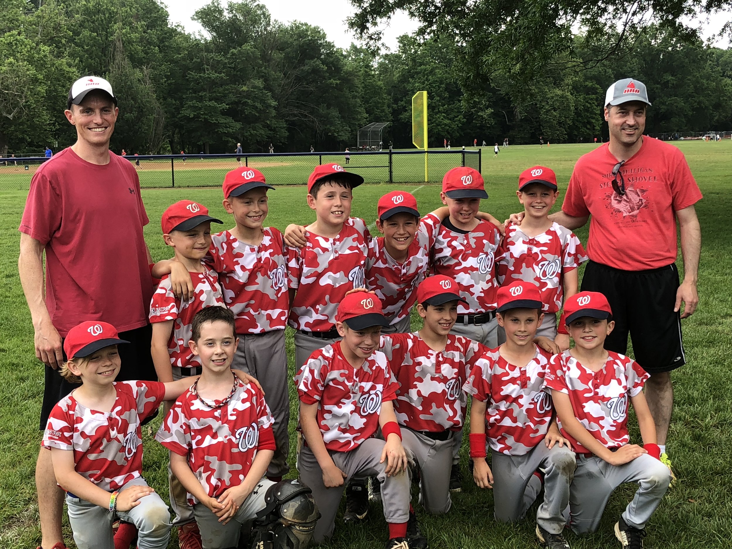 A youth baseball team in red camo jerseys and red caps posing with two adult coaches in red shirts on a baseball field.