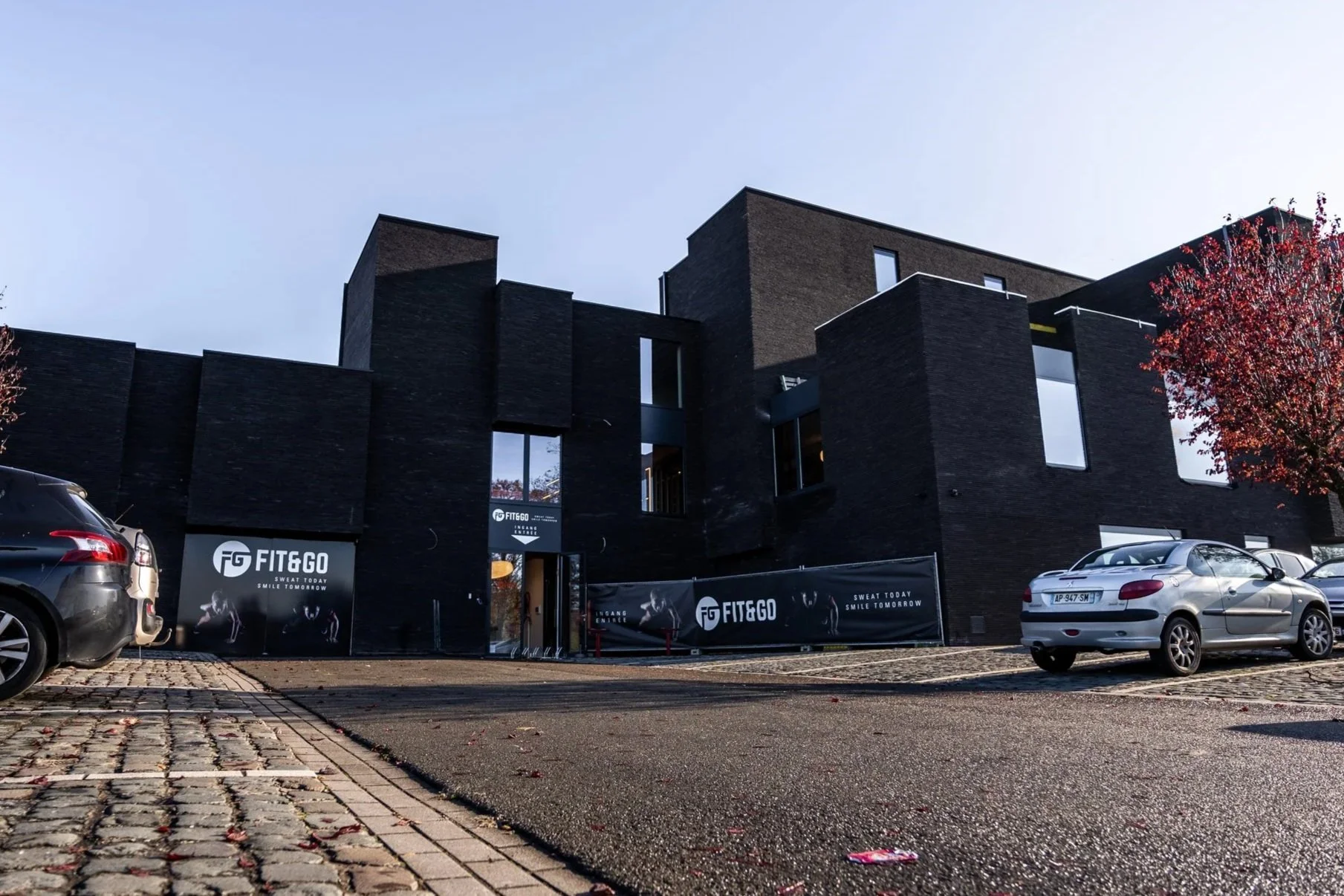 Een zwart modern gebouw met de tekst 'FIT&GO' op banners buiten, parkeervakken en auto’s op de voorgrond, en een rode boom links naast het gebouw, onder een heldere blauwe hemel.