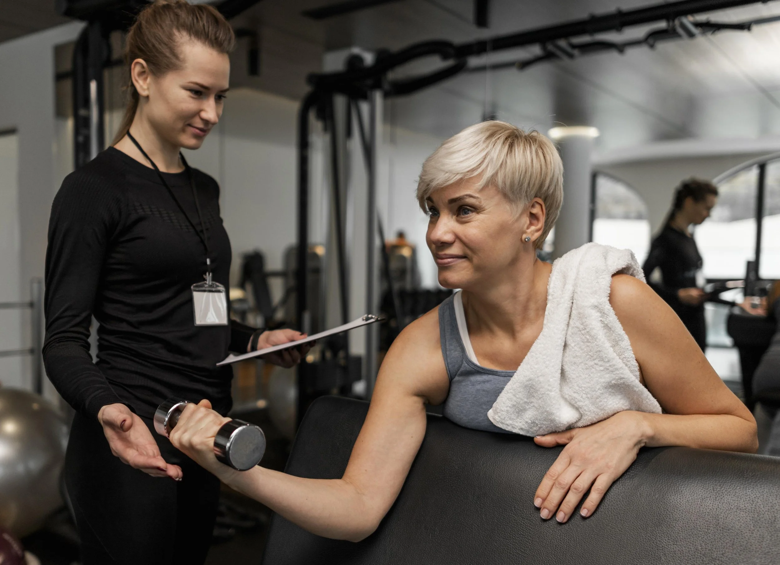 Een vrouw in sportkleding met een handdoek op haar schouder traint met een lichte dumbbell, terwijl een jonge vrouw haar begeleidt en aantekeningen maakt in een sportschool.