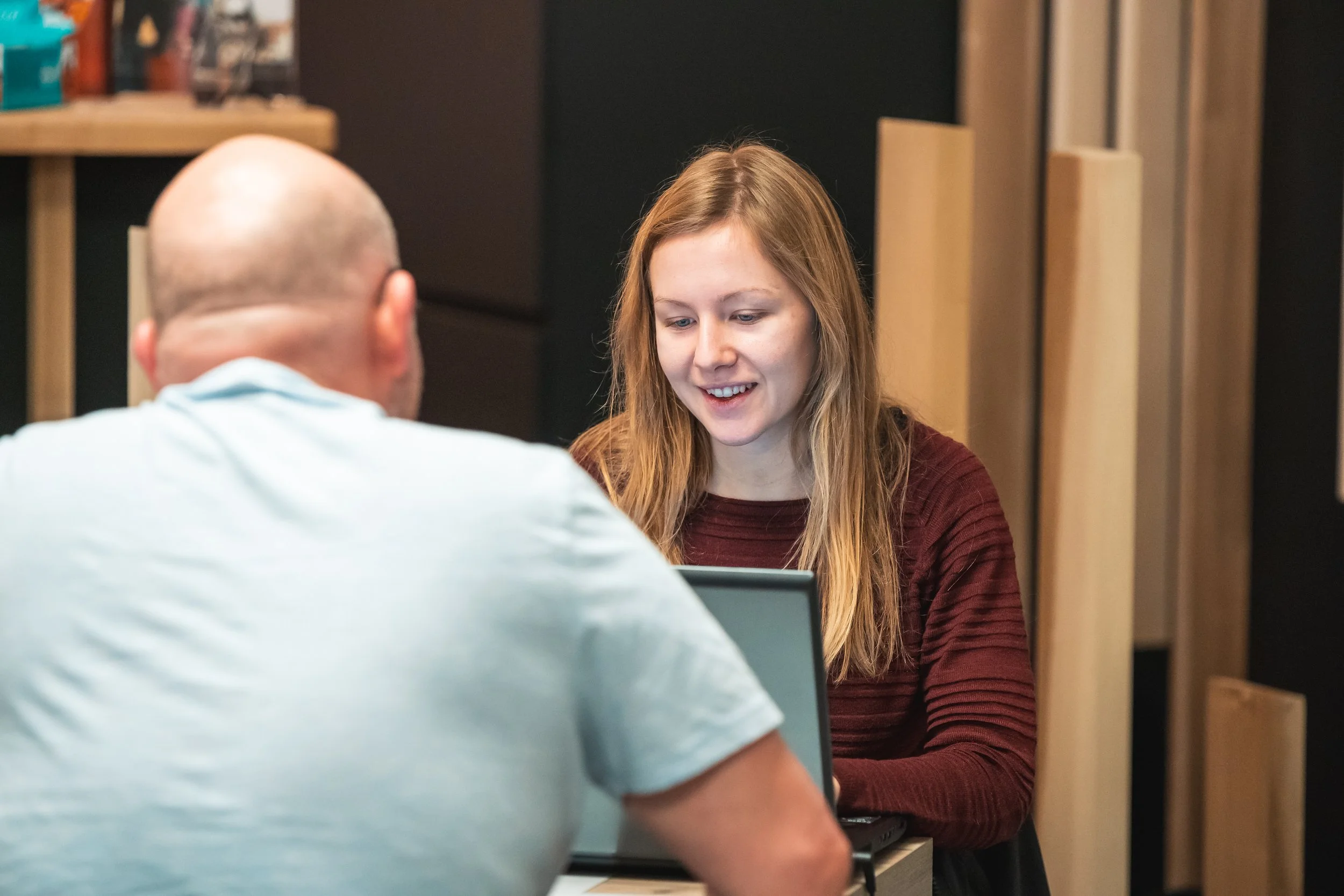 Twee mensen zitten aan een tafel en praten met elkaar, een vrouw met lang blond haar en een man met een kaal hoofd, terwijl ze naar een laptop kijken.