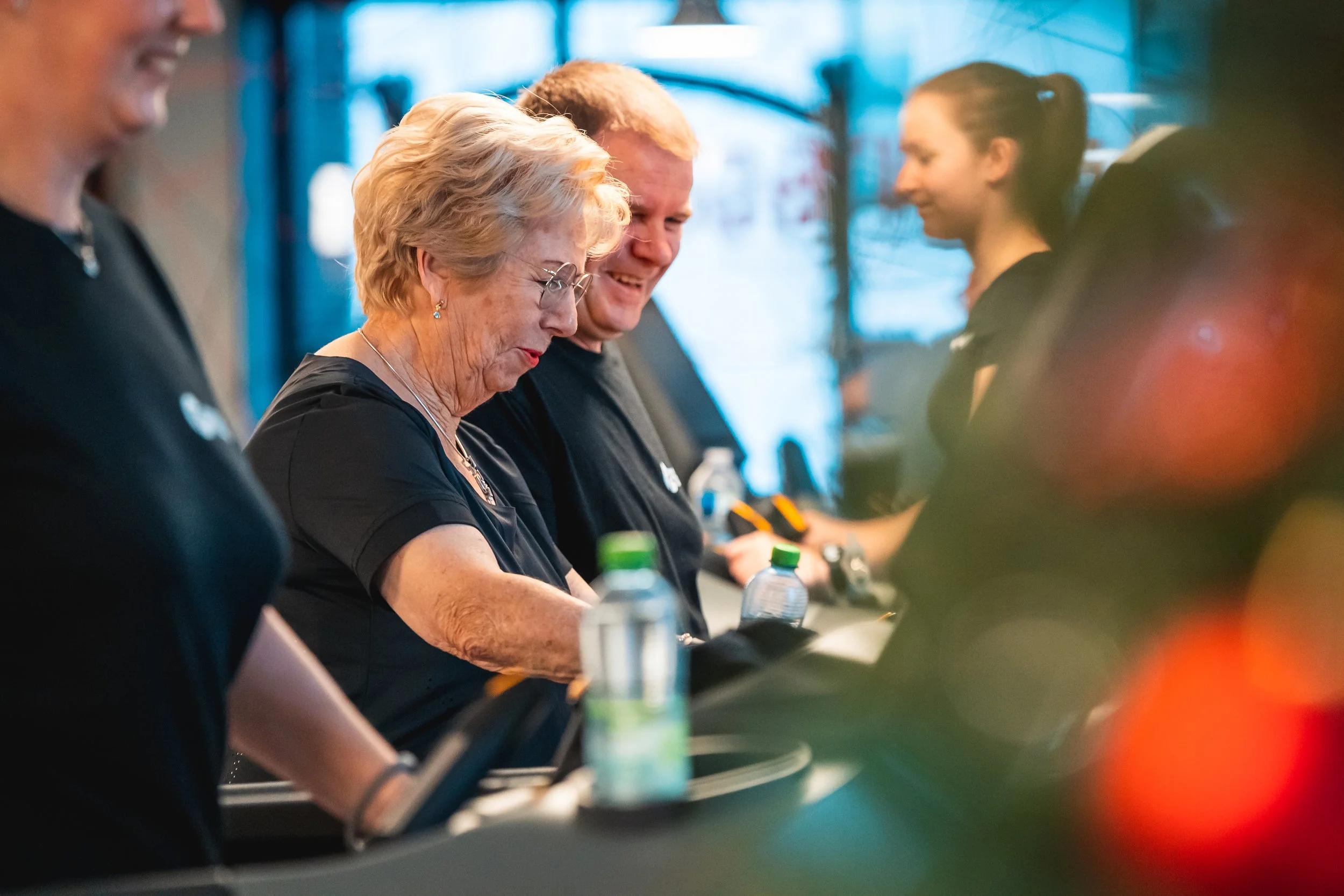 Vijf mensen staan achter een balie, ze dragen zwarte T-shirts en zijn bezig met enkele apparaten. Een oudere vrouw met blonde haar en een bril glimlacht. Een jonge vrouw met een paardenstaart kijkt naar de overkant.