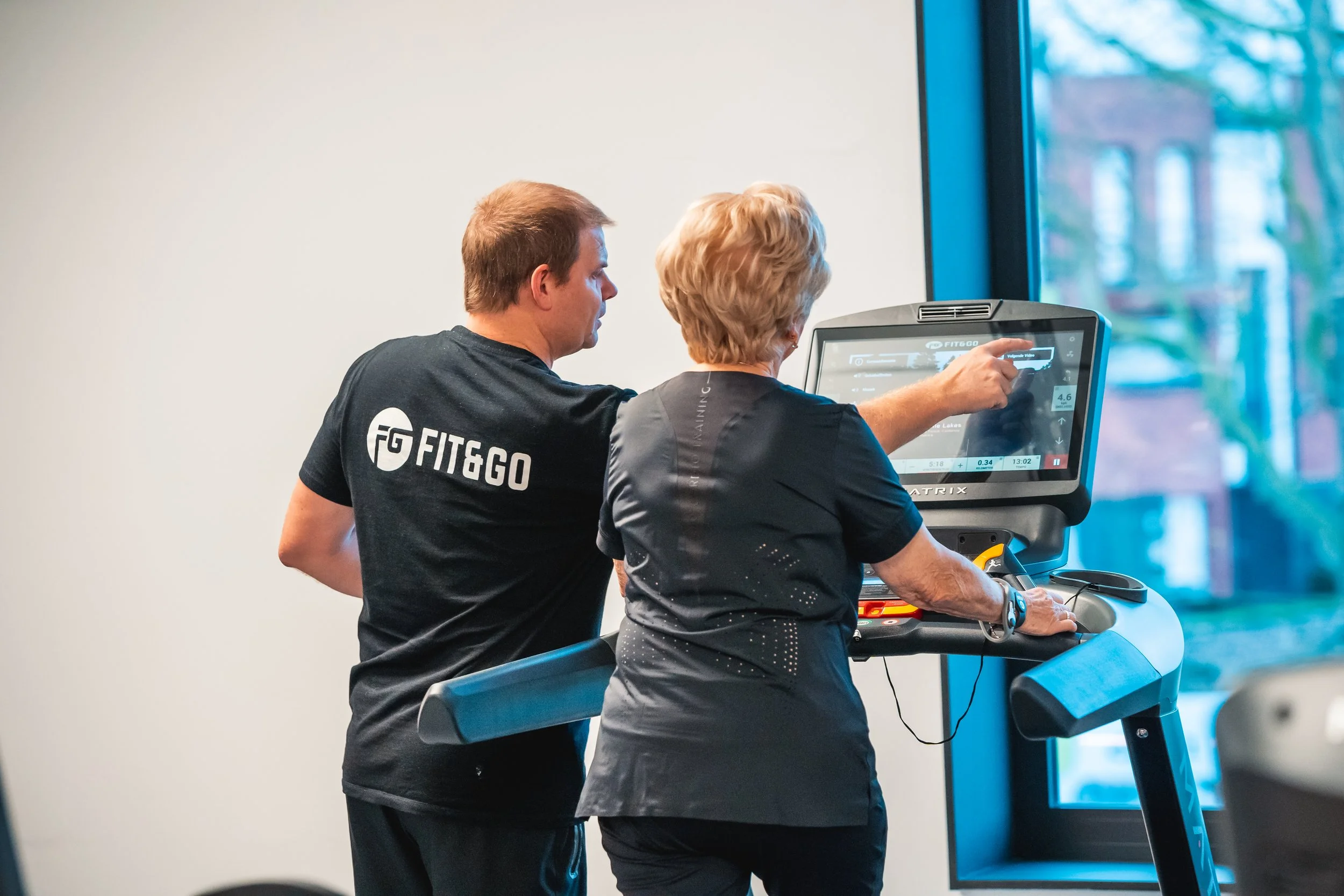 Persoon die in een sportschool op een loopband rent terwijl een trainer instructies geeft met een touchscreen apparaat.
