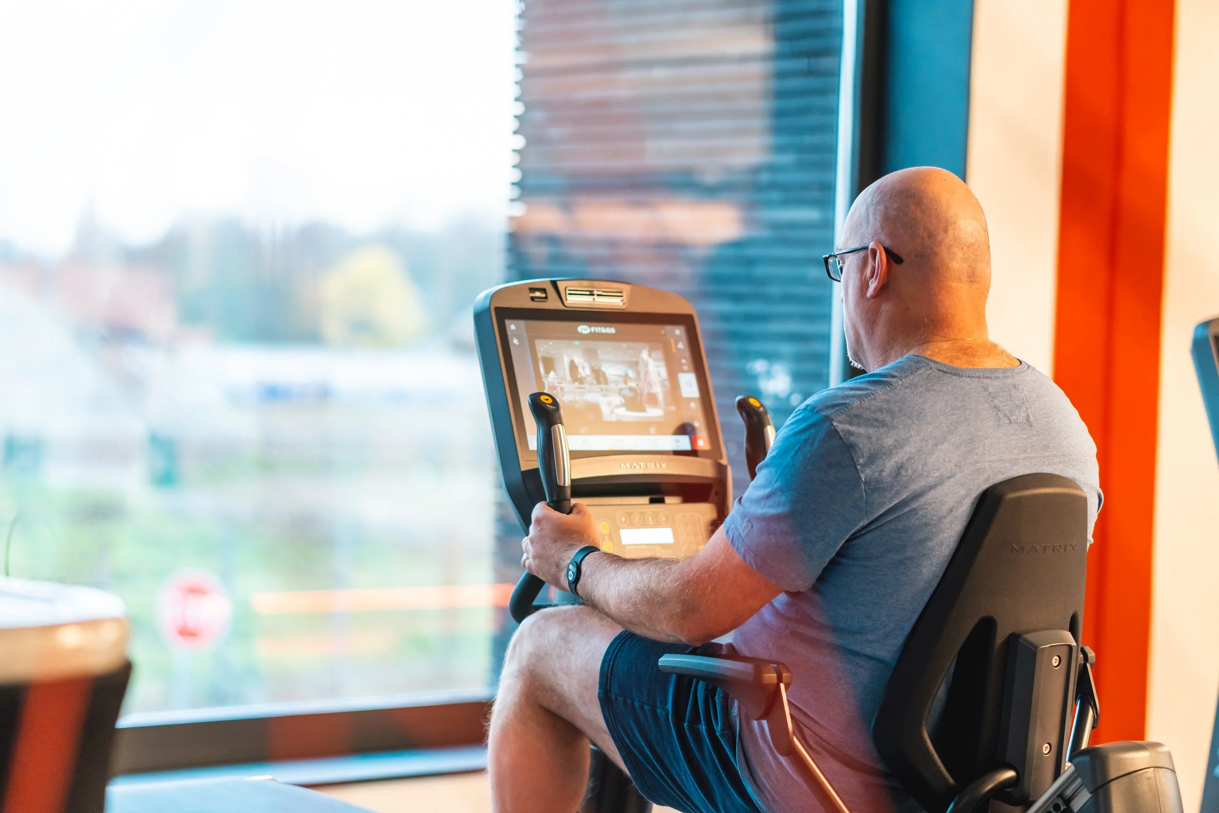 Man zit op een fitnessapparaat en kijkt naar een scherm, terwijl hij binnen een sportschool zit.