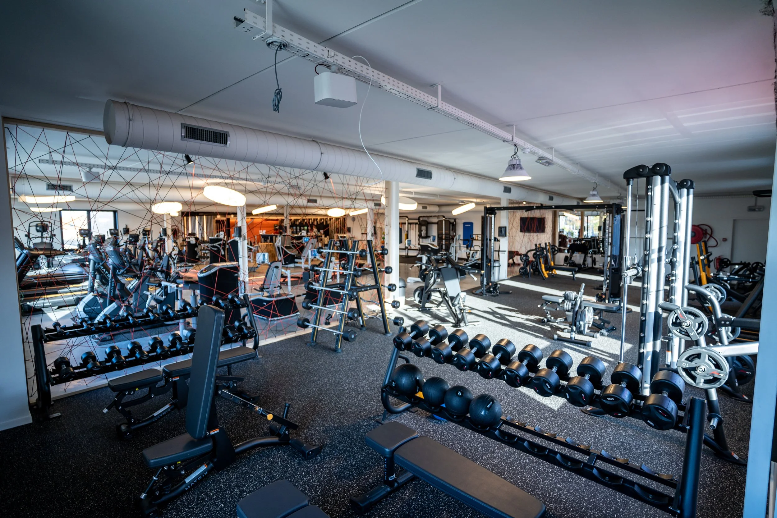 Binnenkant van een sportschool met verschillende mensen die cardio-apparatuur gebruiken en fitnessapparatuur zoals gewichten en machines.