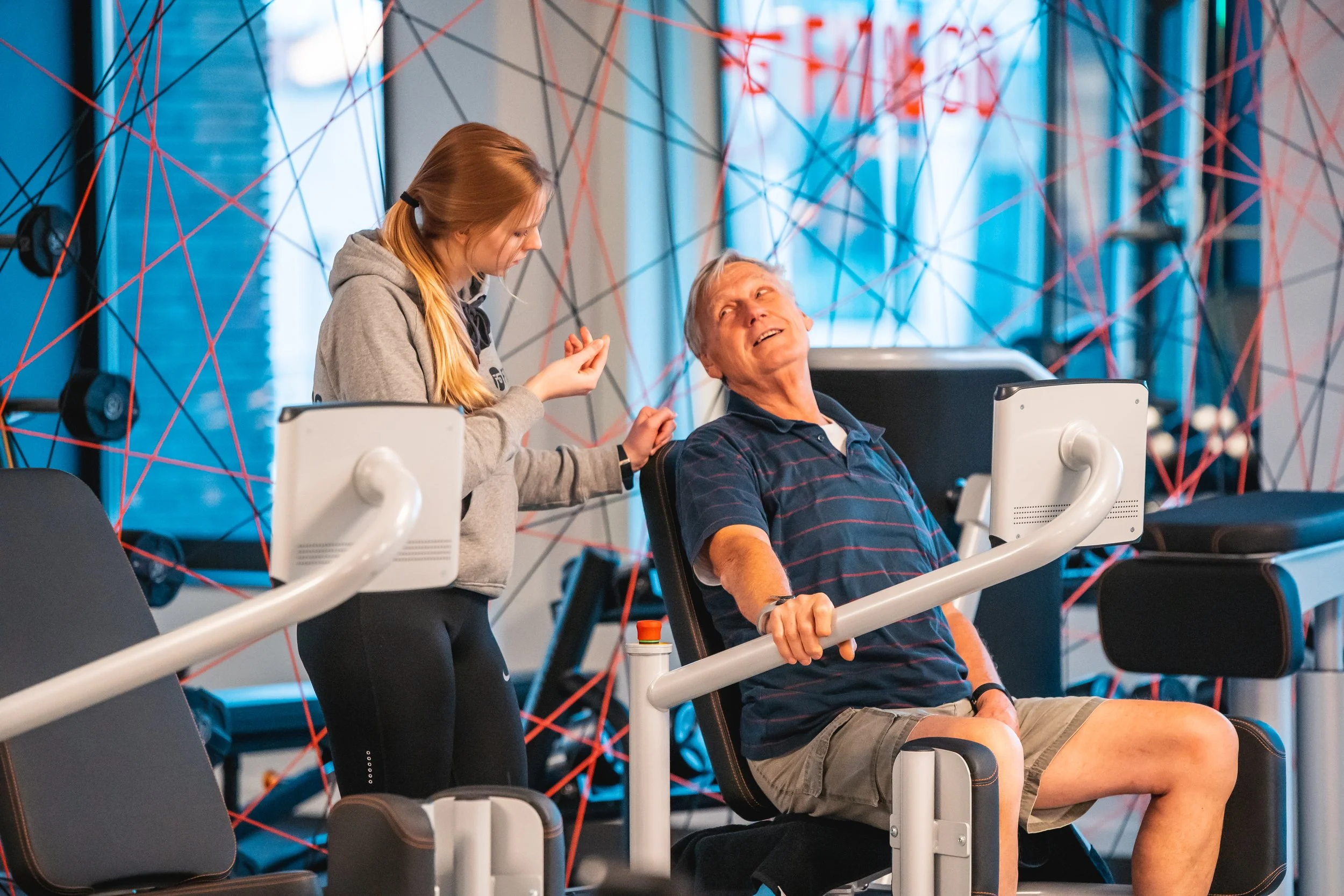 Een jonge vrouw die aan het trainen is in een sportschool met een oudere man die op een fitnessapparaat zit en lacht.