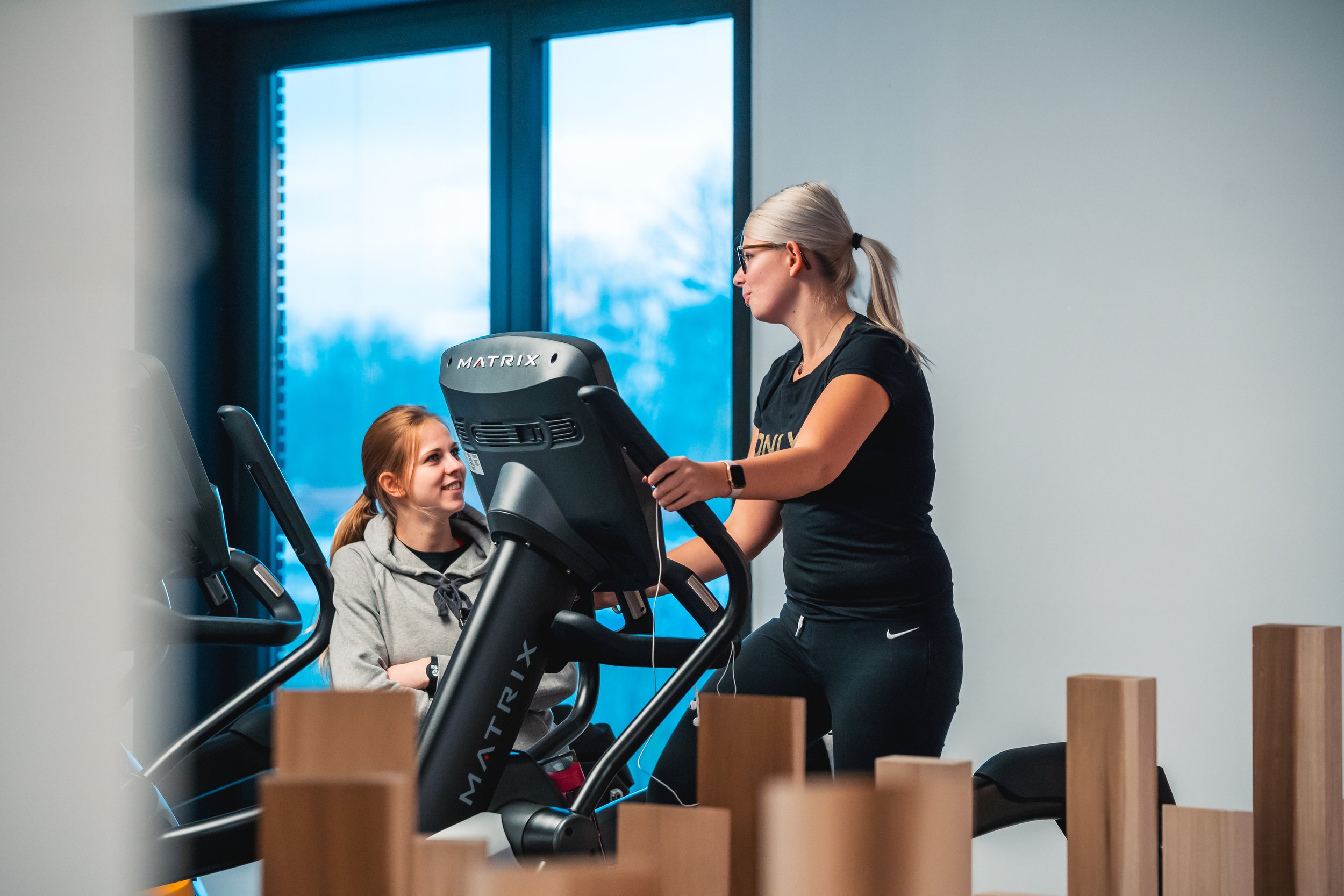 Een vrouw die op een loopband traint met een glimlach, terwijl een trainster of begeleider naast haar staat en haar aanmoedigt in een fitnessruimte.