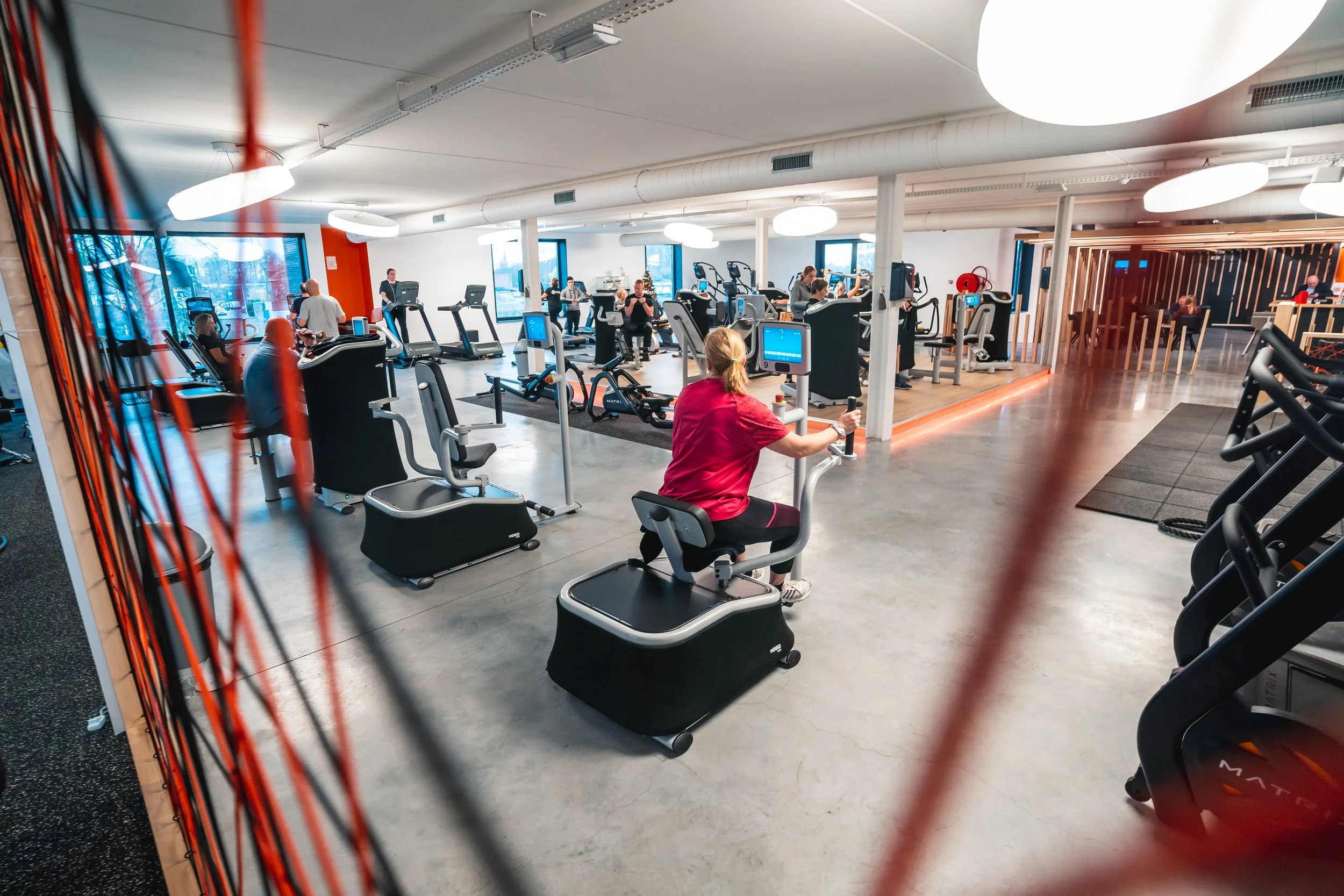 Binnen een fitnesscentrum met diverse mensen die apparaten gebruiken voor cardio- en krachttraining, met grote ramen en moderne verlichting.