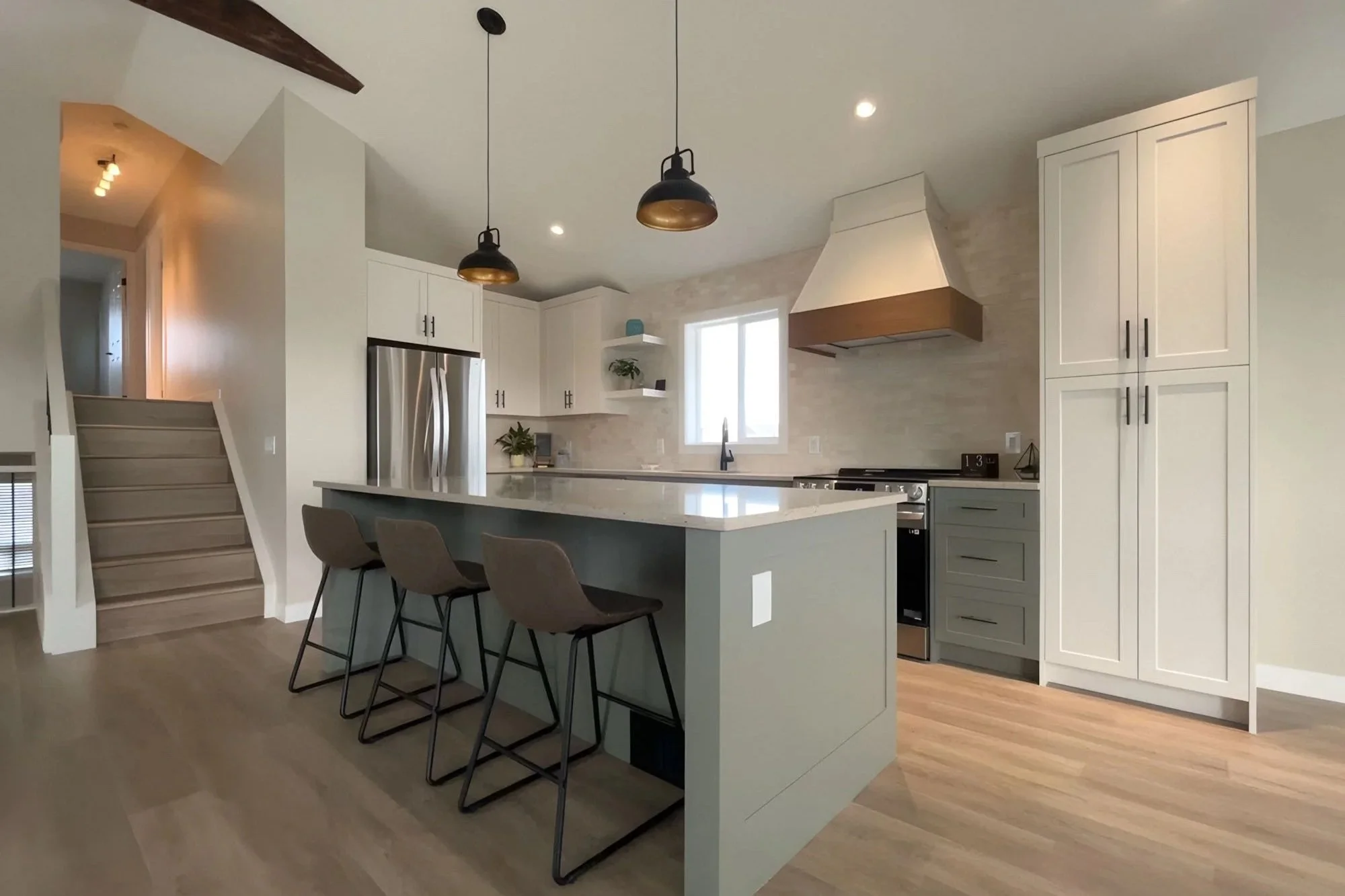 Modern kitchen showcasing custom residential carpentry. Cabinets, stainless steel refrigerator, island with gray base and three chairs, black pendant lights, window above sink, and wooden flooring.