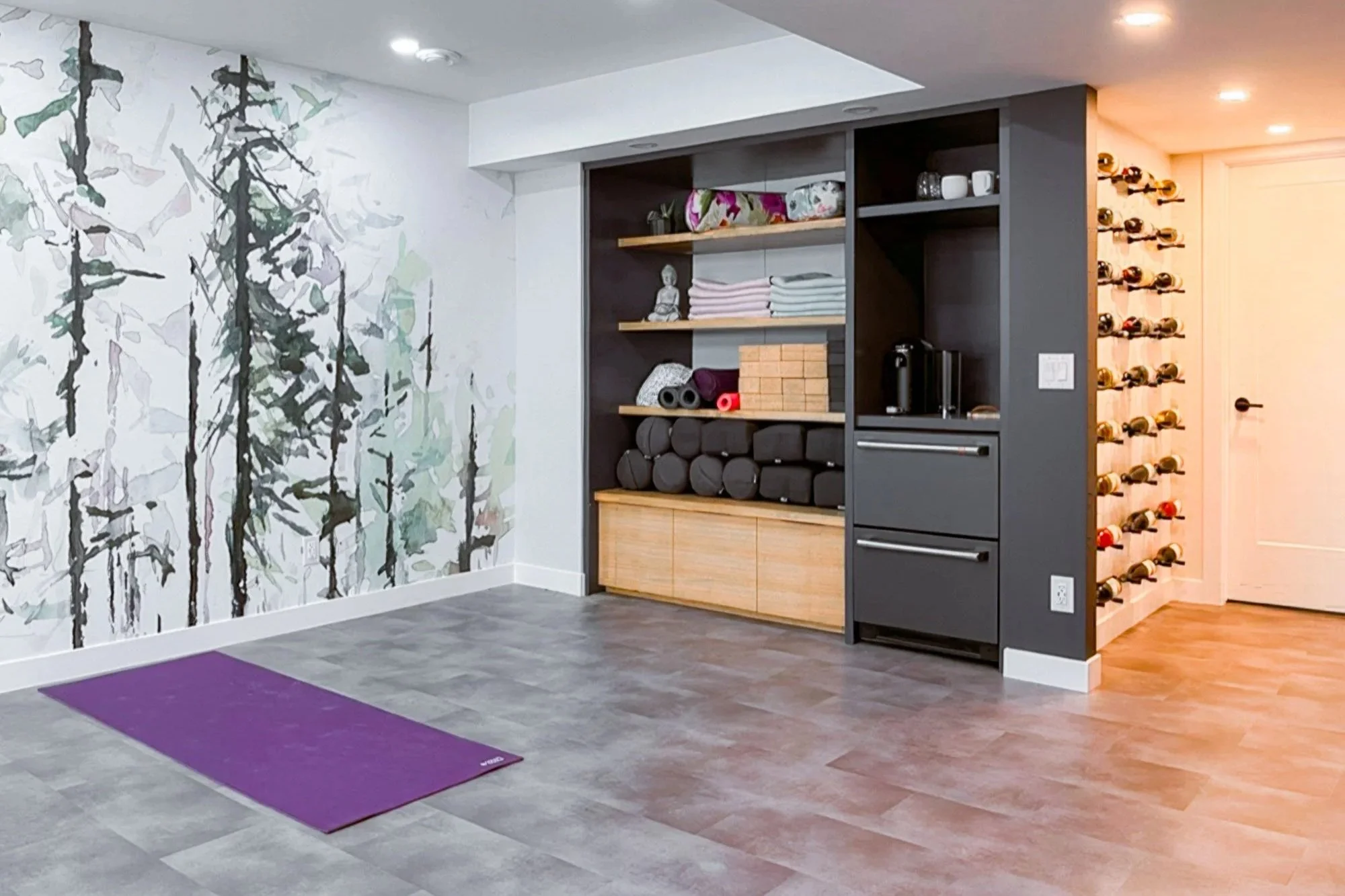 A basement renovation featuring a home yoga studio with a purple mat on the floor, a large forest-themed mural on the wall. To the right, there's built-in cabinetry and a custom wine rack display unit.
