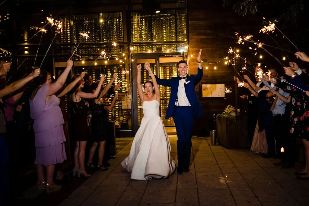 A bride and groom walking hand in hand during their wedding celebration, surrounded by guests holding sparklers at night.