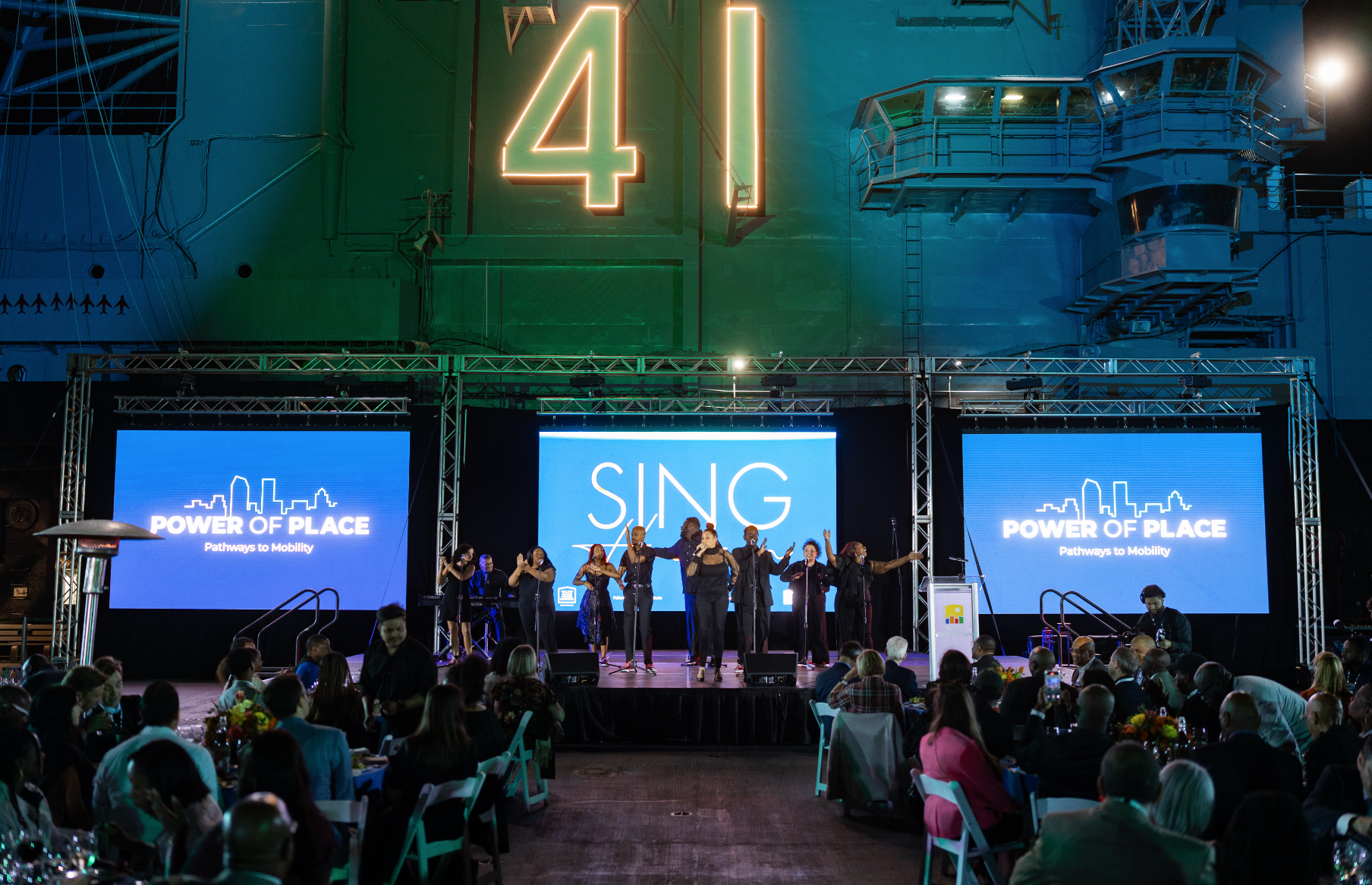 A stage with singers performing at an event titled 'Power of Place: Pathways to Mobility,' with large screens displaying the event theme, and a crowd seated at tables in front of the stage, in a large industrial building.