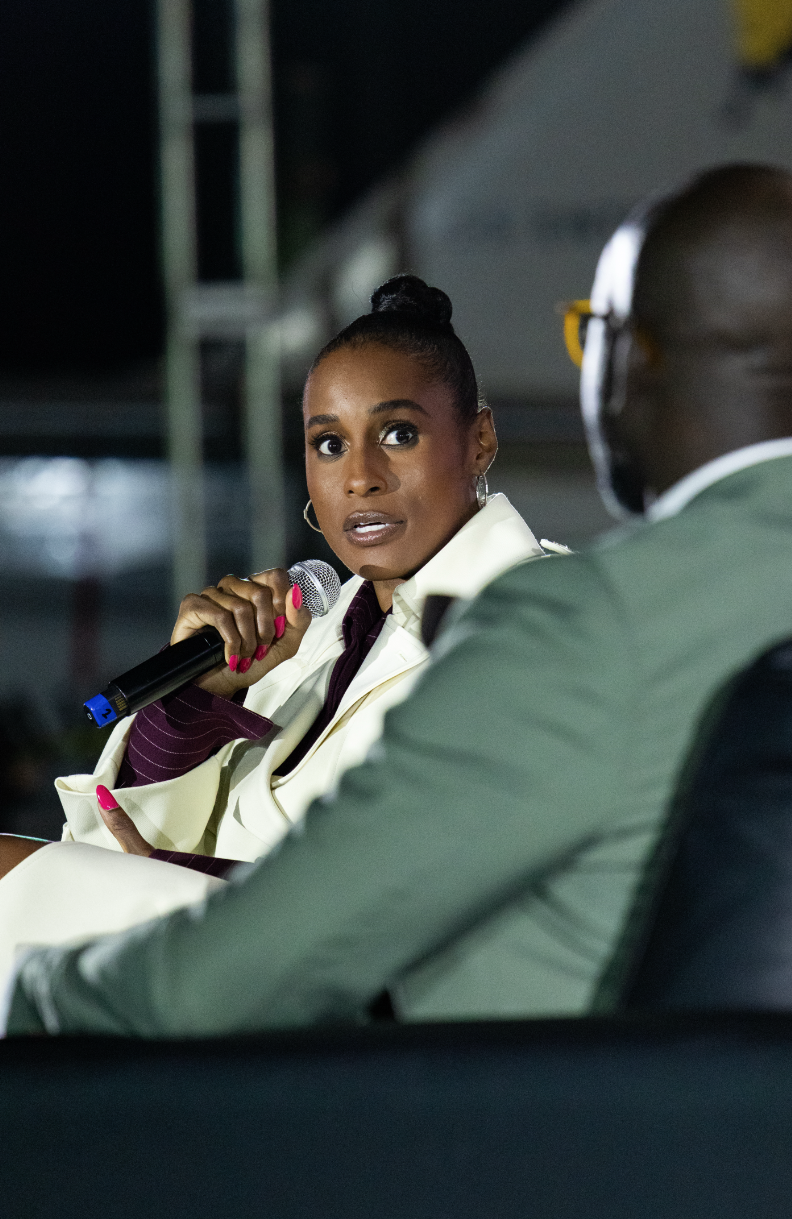 A woman holding a microphone appears to be speaking or asking a question during an event, as she looks intently at a man seated across from her, in an indoor setting with a dark background.