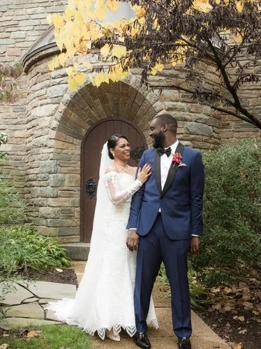 A bride and groom stand together outdoors in front of a stone building, smiling at each other. The bride is in a white lace wedding dress with long sleeves, and the groom is in a navy suit with a white shirt and black bow tie, holding hands. There are green bushes and a tree with yellow leaves nearby.