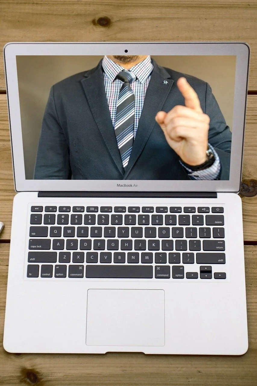 The image depicts a MacBook Air laptop on a wooden surface, with the laptop's screen showing a person in a business suit pointing one finger upward. The person is dressed in a dark suit, checkered shirt, and striped tie.