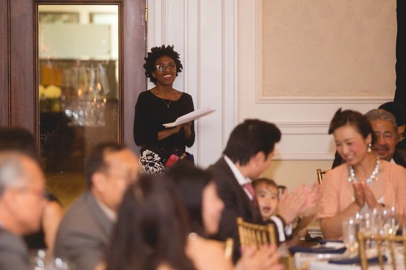 A woman standing and holding papers, smiling at a formal event with seated guests clapping and smiling.