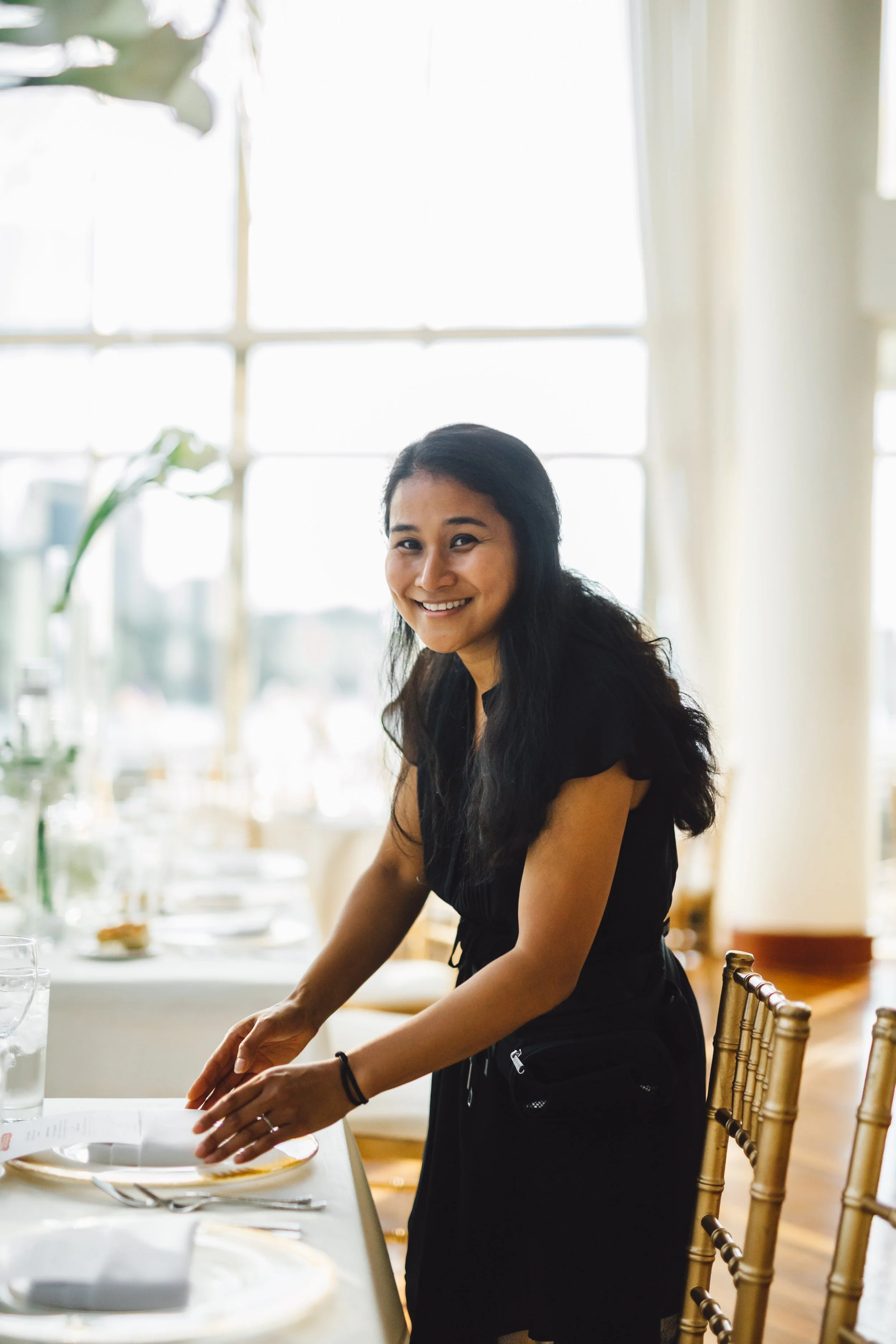 A woman with long dark hair wearing a black dress, smiling and setting a table in a brightly lit room with large windows and elegant decor.