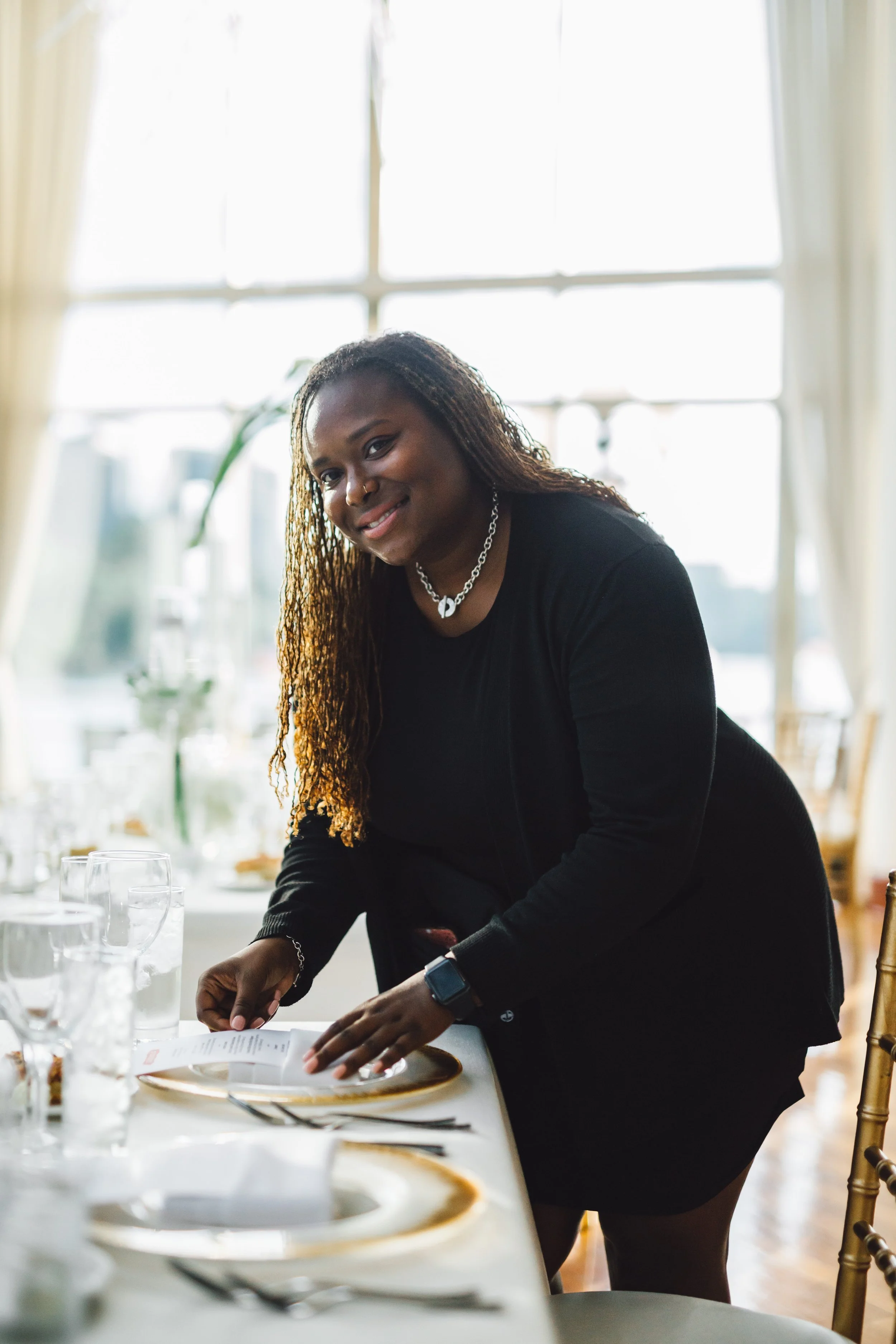 A woman setting a dining table in a bright room with large windows, wearing a black dress, silver jewelry, and a smartwatch.