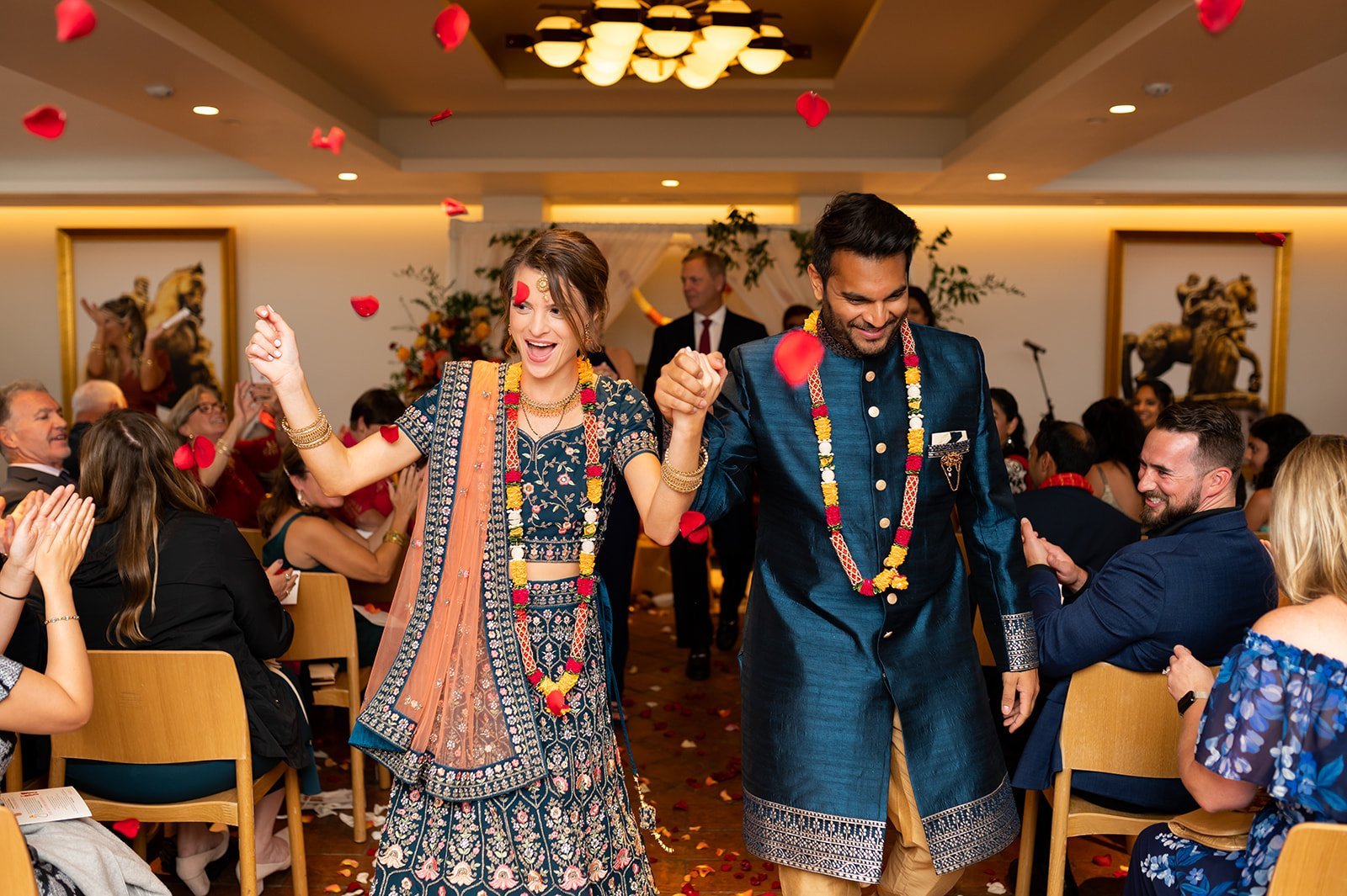 A woman and a man dressed in traditional Indian attire celebrating during a wedding ceremony, holding hands and smiling amid falling rose petals.