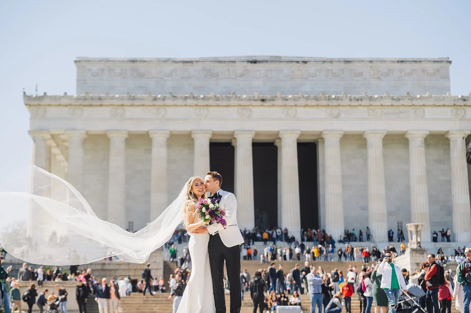 A bride and groom standing in front of the Lincoln Memorial in Washington, D.C. The bride is holding a bouquet of purple and white flowers, and her veil is flowing in the wind. There are many people in the background