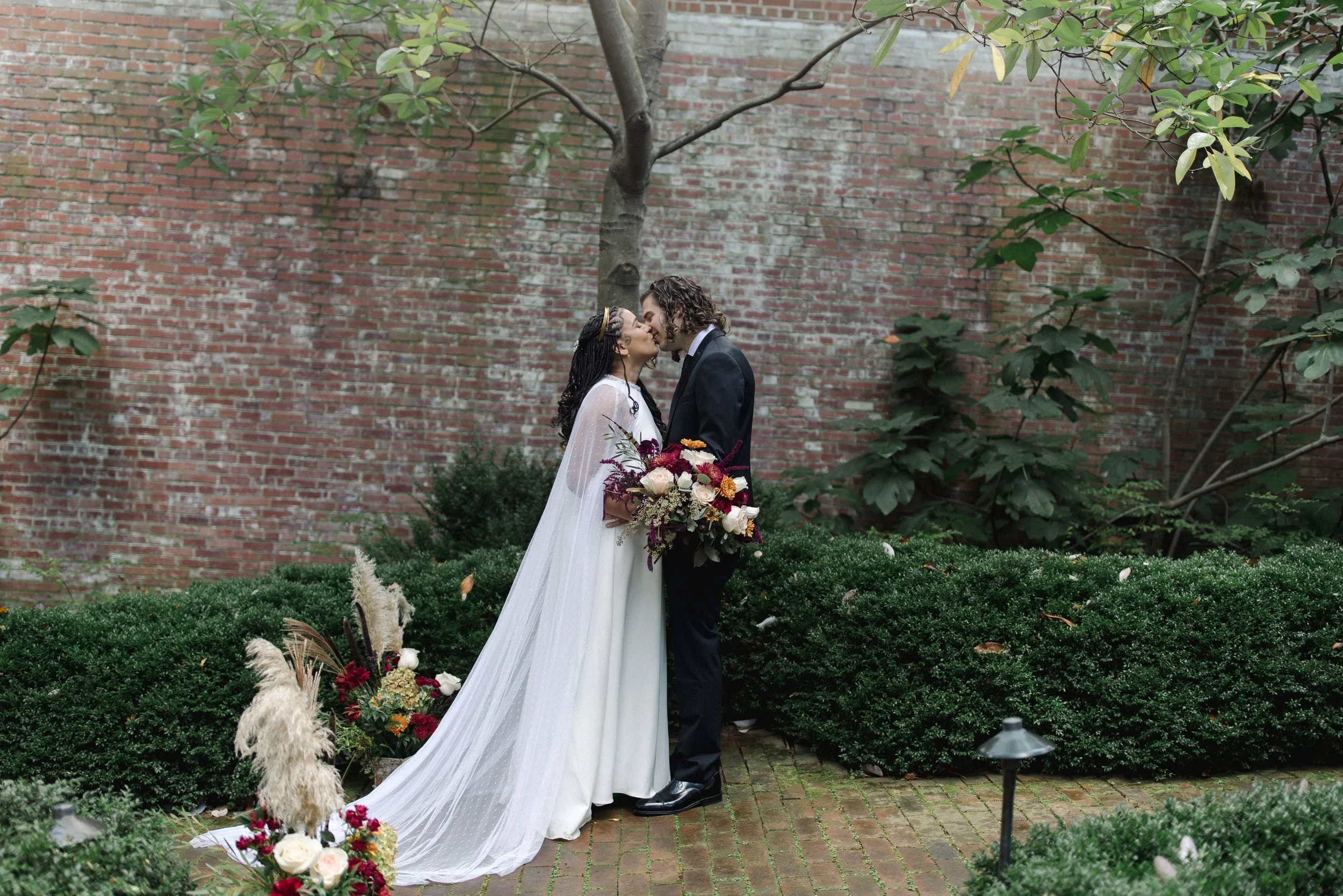A bride and groom kiss in an outdoor garden with a brick wall background. The bride wears a white gown with a long train and holds a bouquet of flowers, while the groom is dressed in a black suit. There are floral arrangements and greenery around the