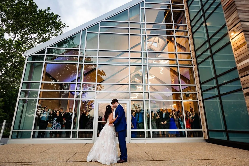 A bride and groom are standing close together, holding hands, outside a modern glass building with a large crowd of wedding guests visible inside behind the glass.